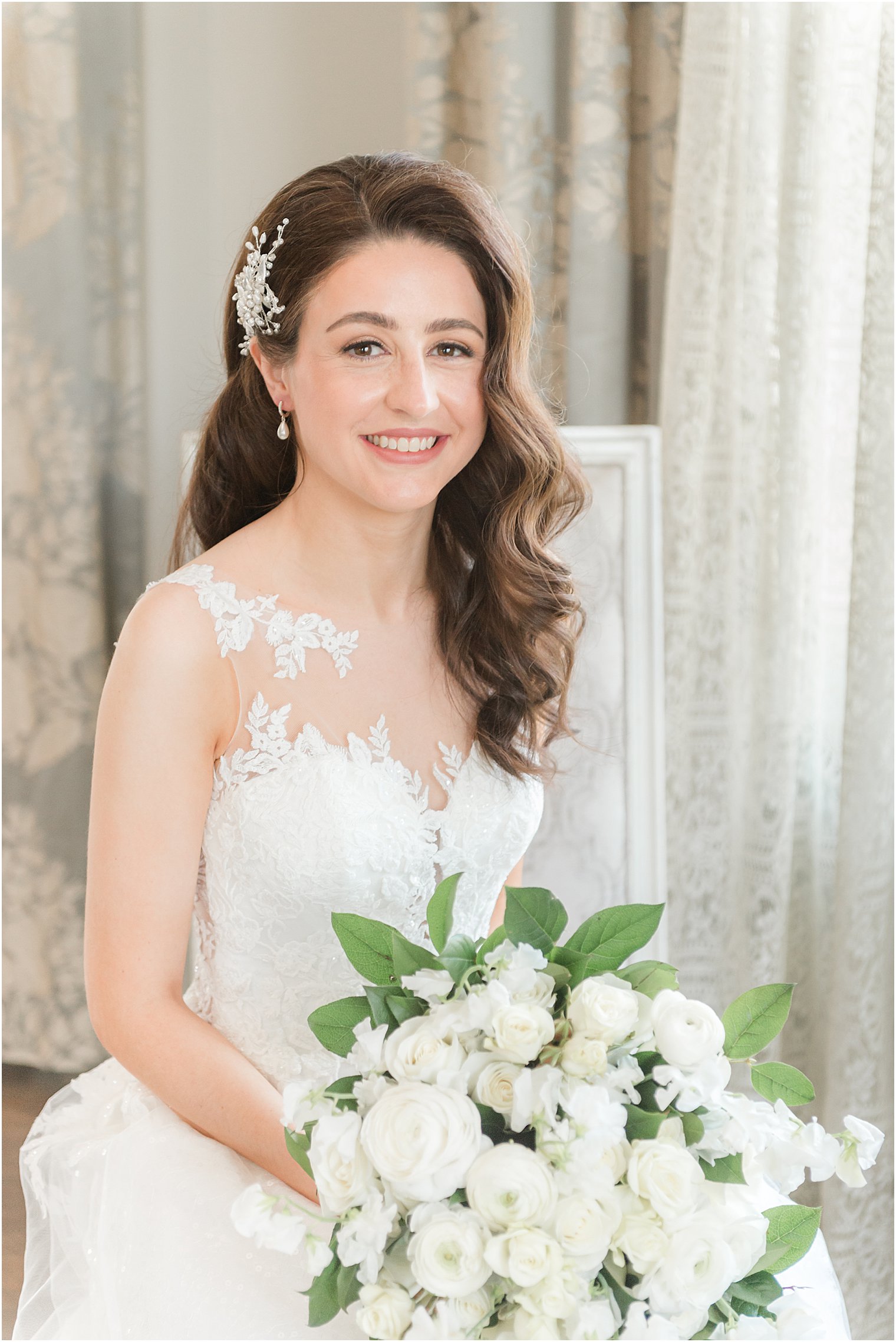 bride sits in chair in wedding gown with bouquet of white roses in lap 