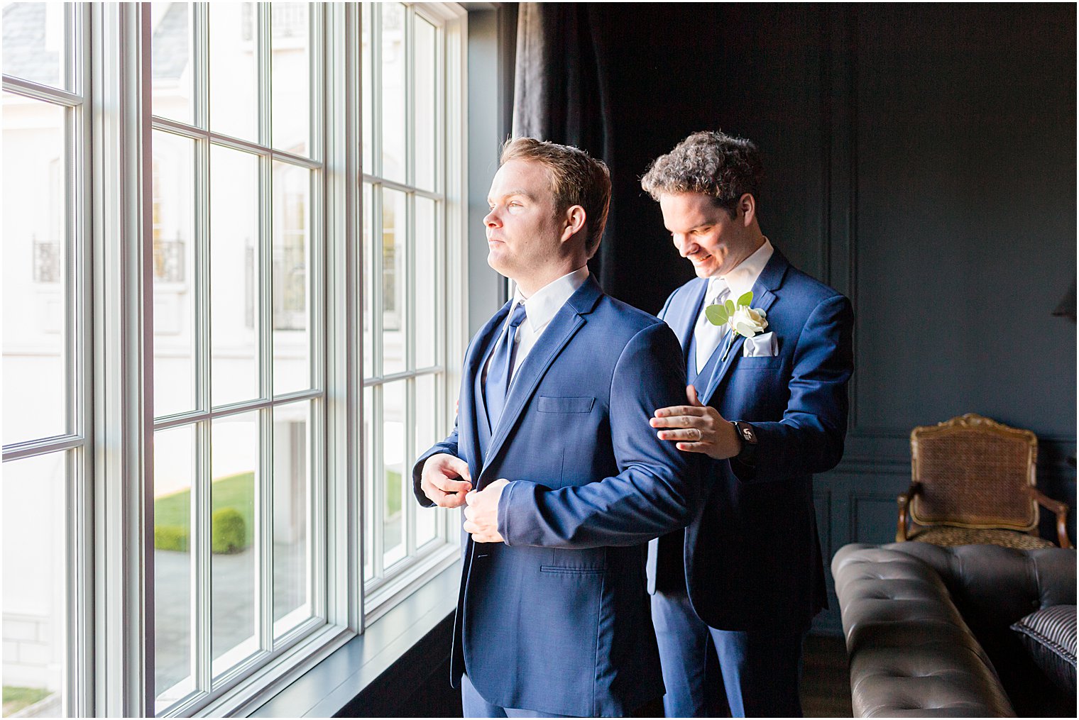 groomsman helps groom with blue suit jacket 