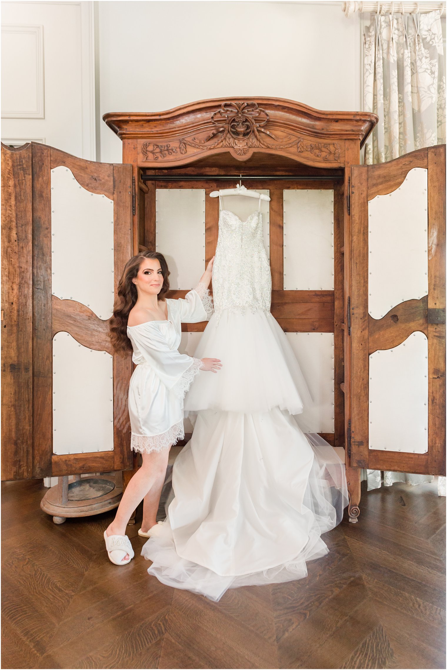 bride in white robe looks at mermaid wedding gown hanging in wooden armoire at Park Chateau Estate
