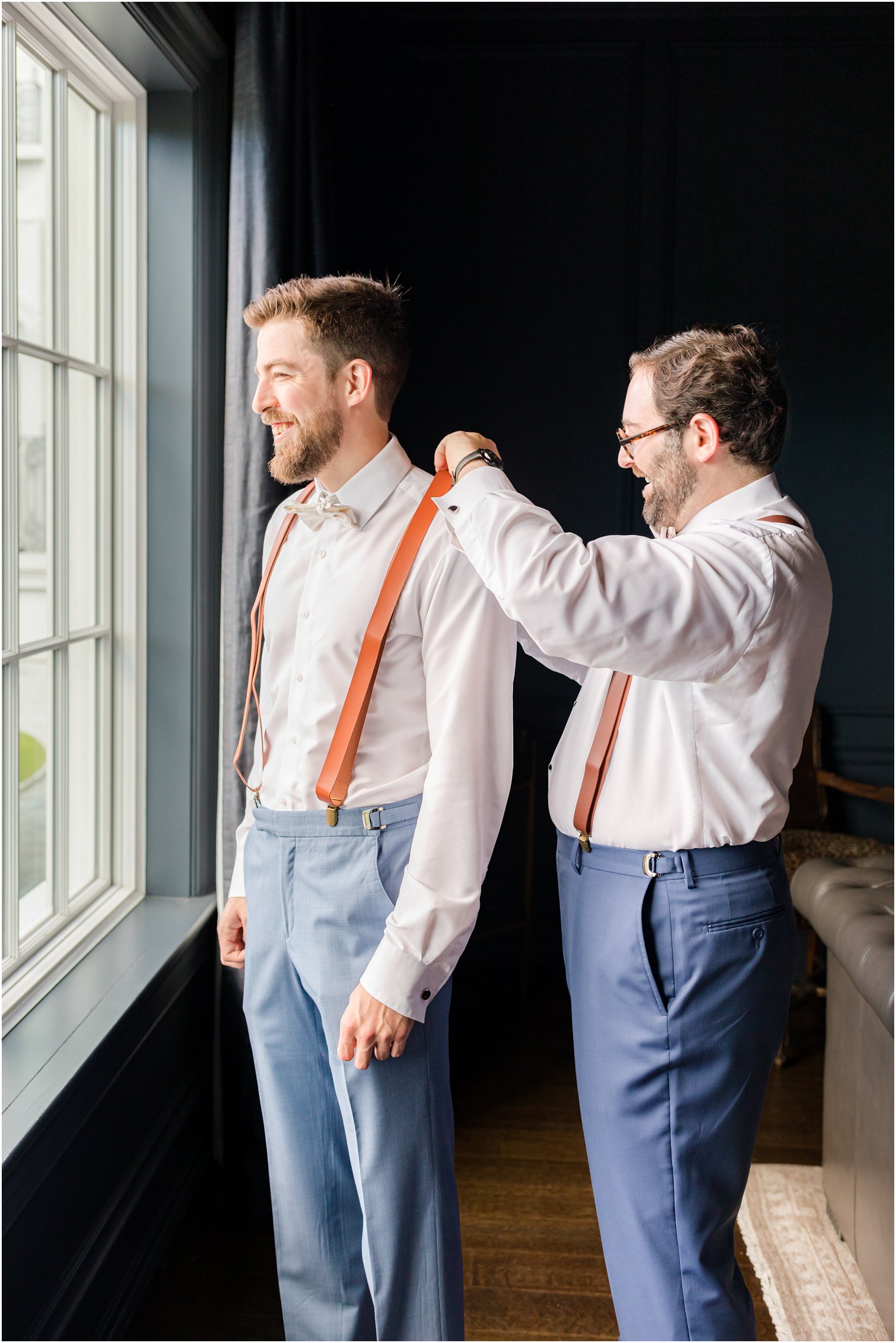 groomsman helps groom with suspenders by window at Park Chateau Estate