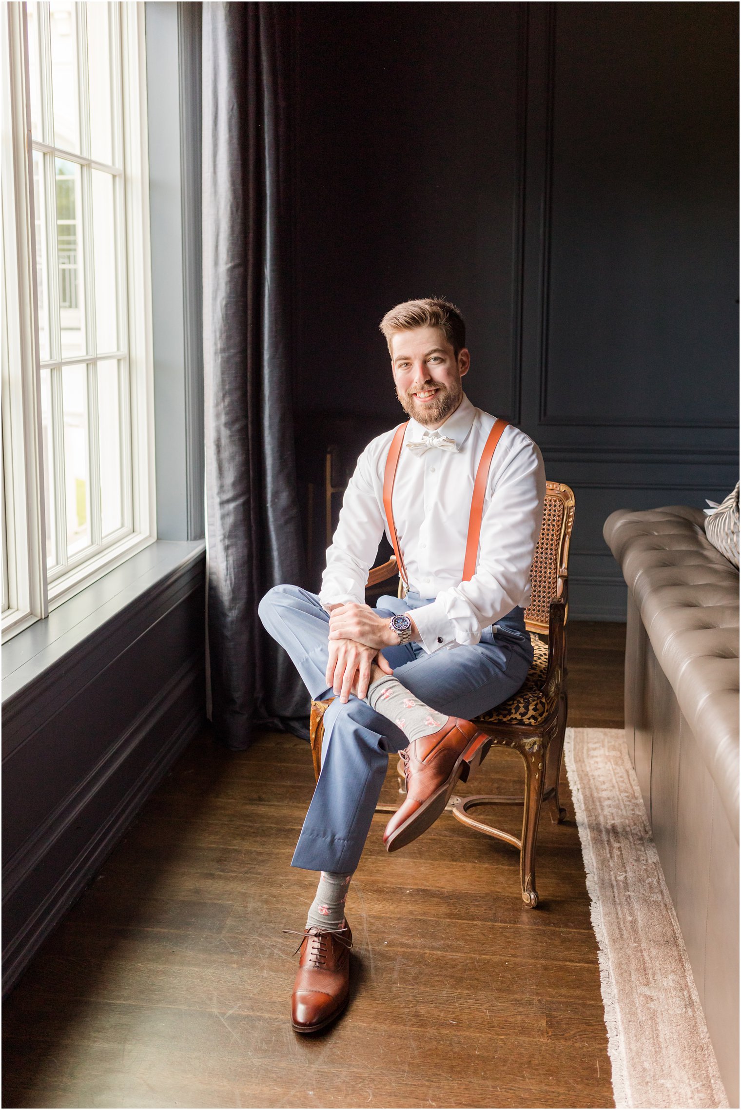 groom sits in wooden chair by window in suspenders and blue pants at Park Chateau Estate
