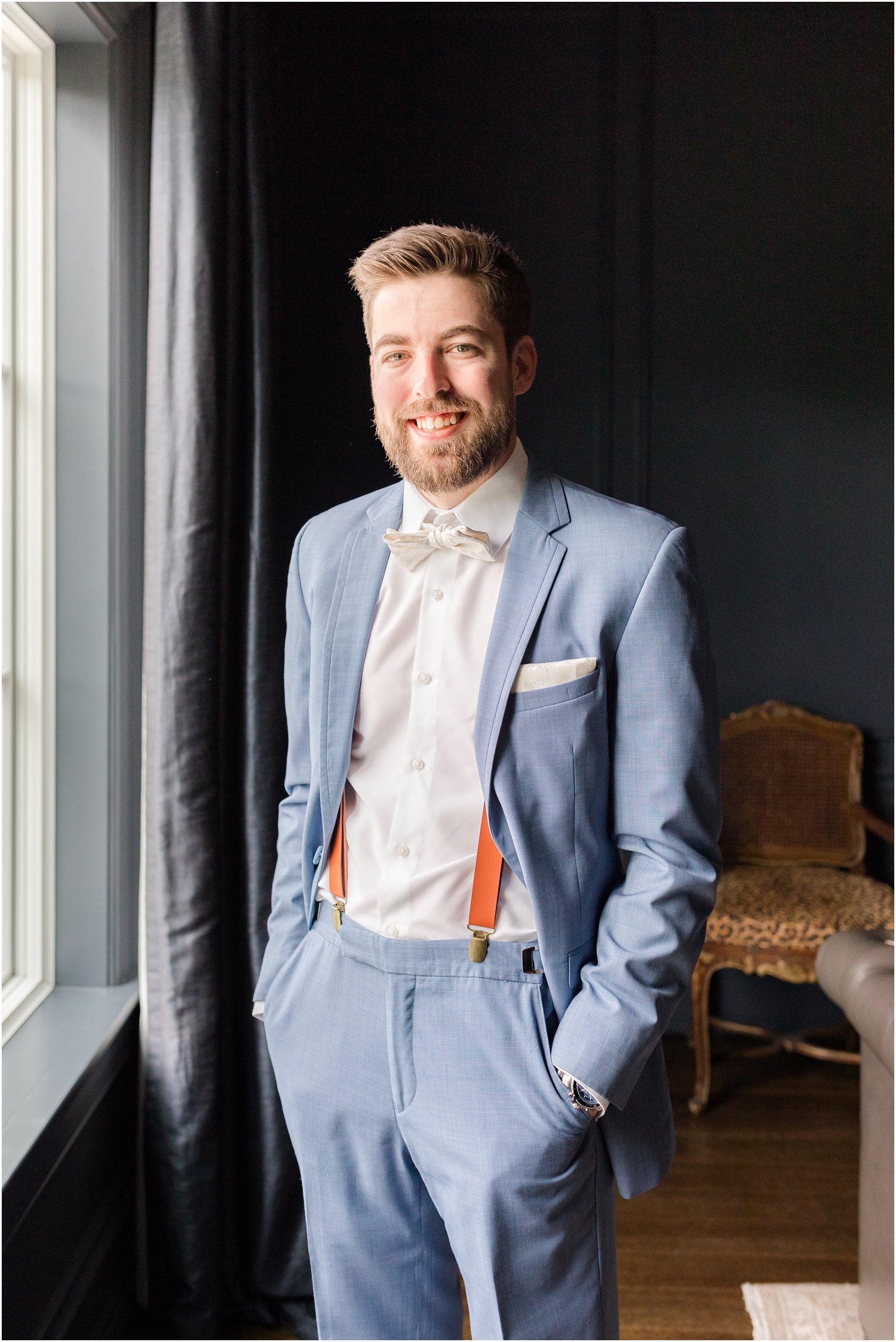 groom stands in light blue suit with hands in pockets by window in suite 