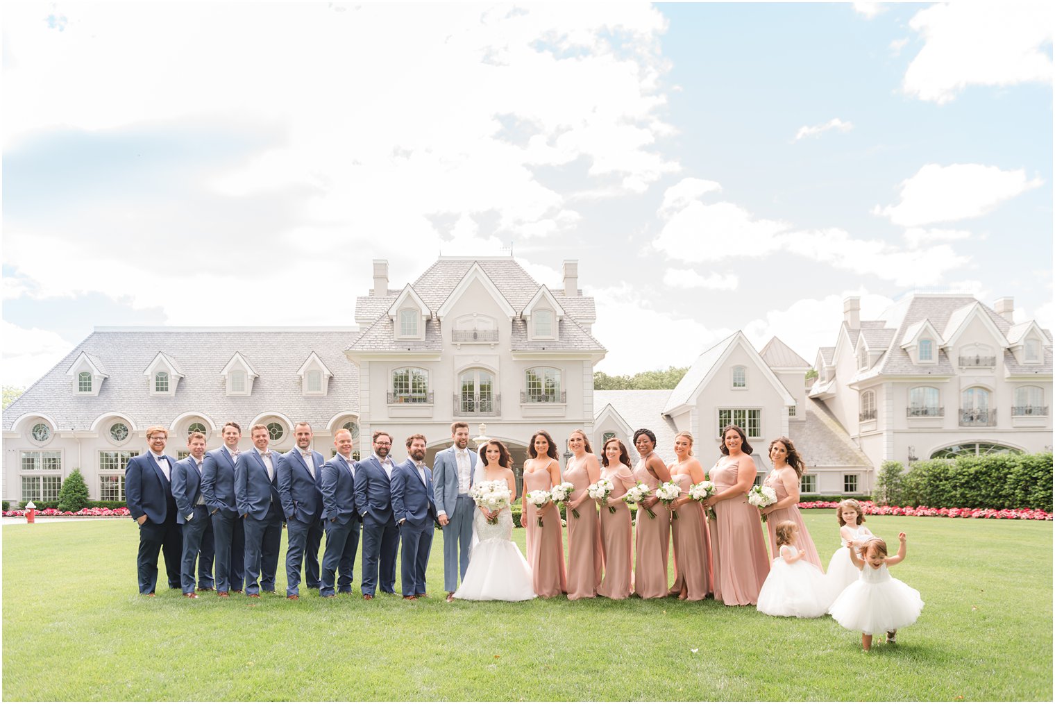 bride and groom stand on lawn with groomsmen in navy suits and bridesmaids in pink gowns on lawn