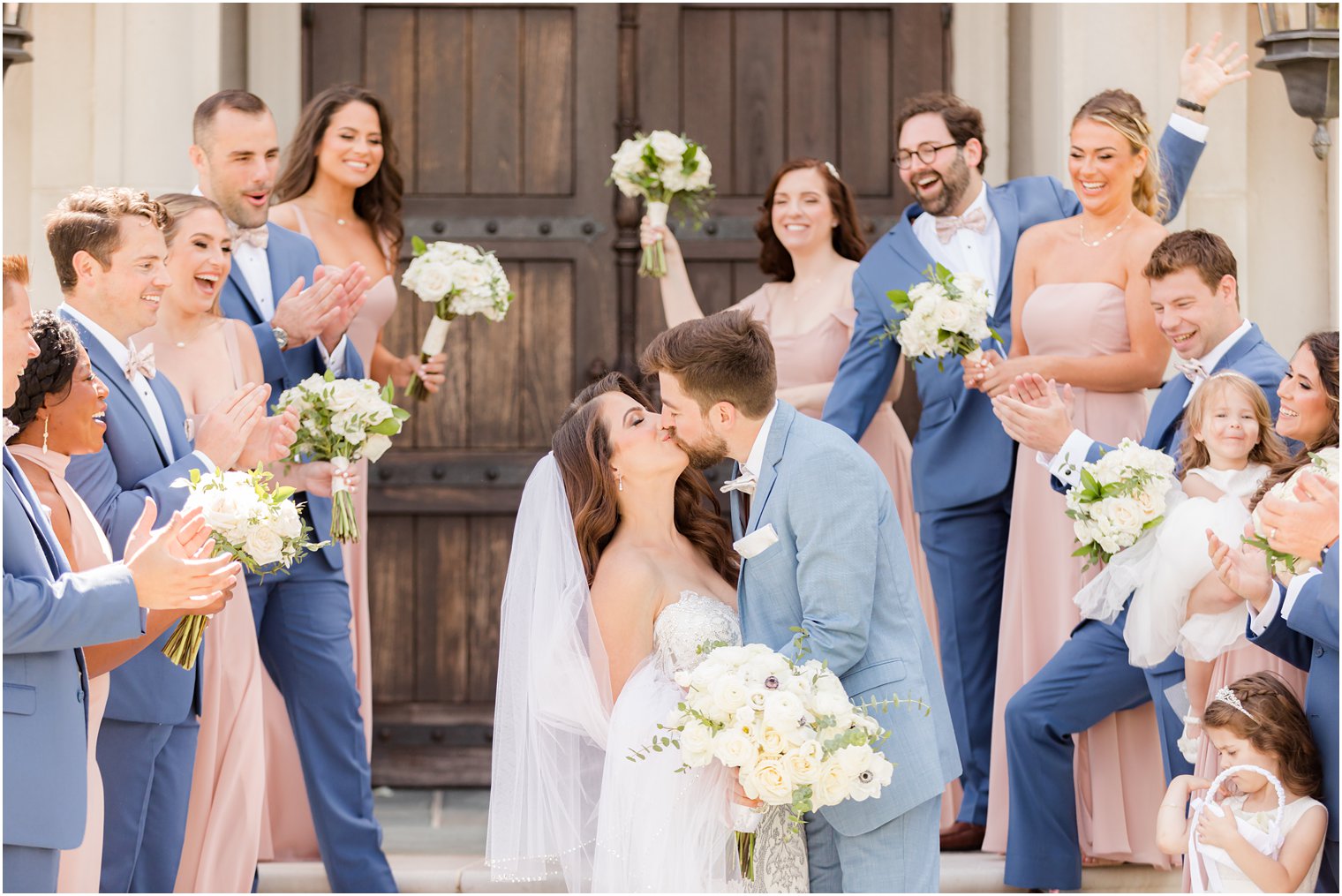 bride and groom kiss ons tips by chapel at Park Chateau near wooden doors with bridal party cheering and clapping around them 
