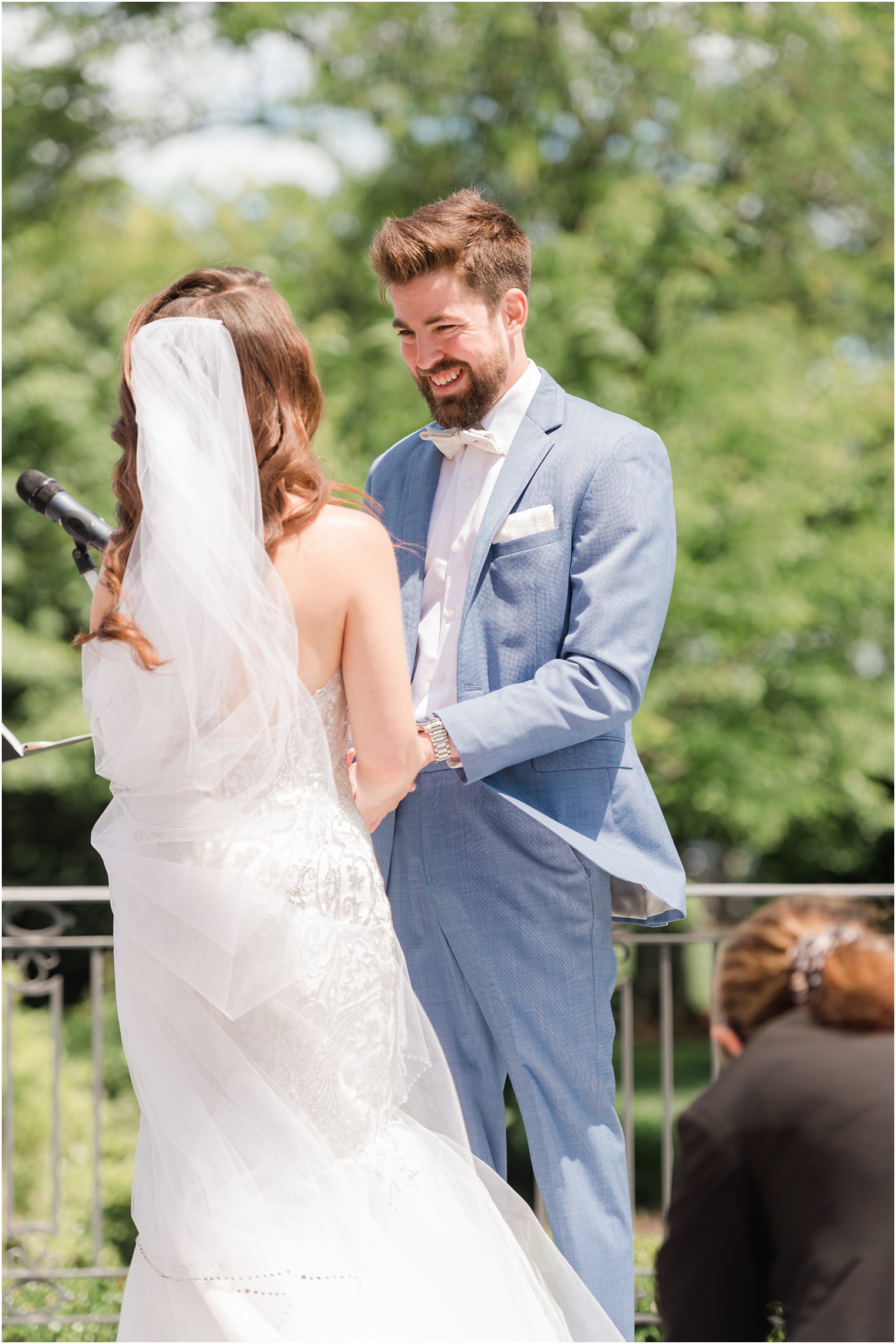 groom in blue suit smiles at bride during windy wedding ceremony in gardens of Park Chateau 