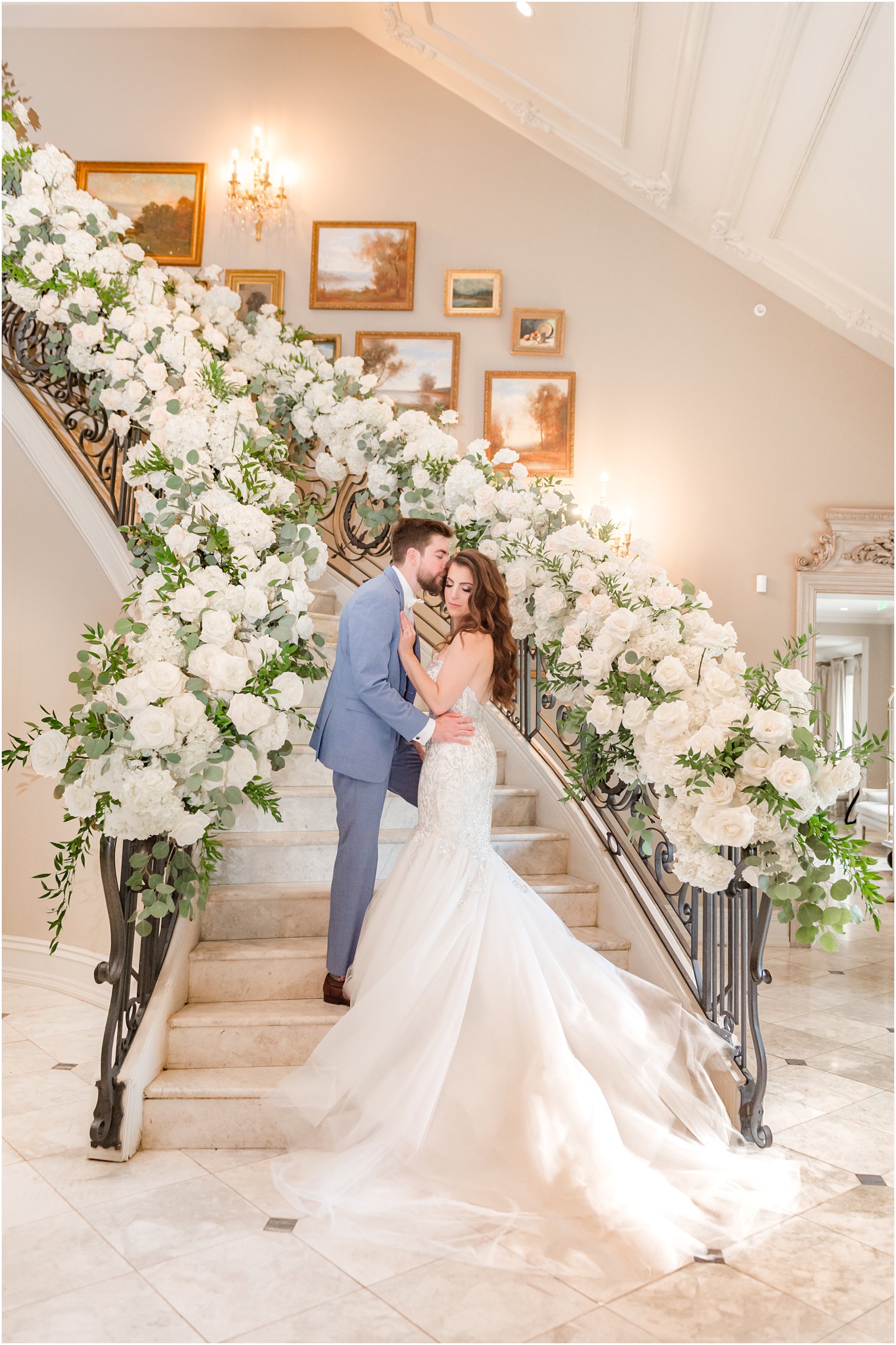 groom in blue suit leans to kiss bride's forehead standing on steps inside Park Chateau Estate with pink rose floral display down staircase 