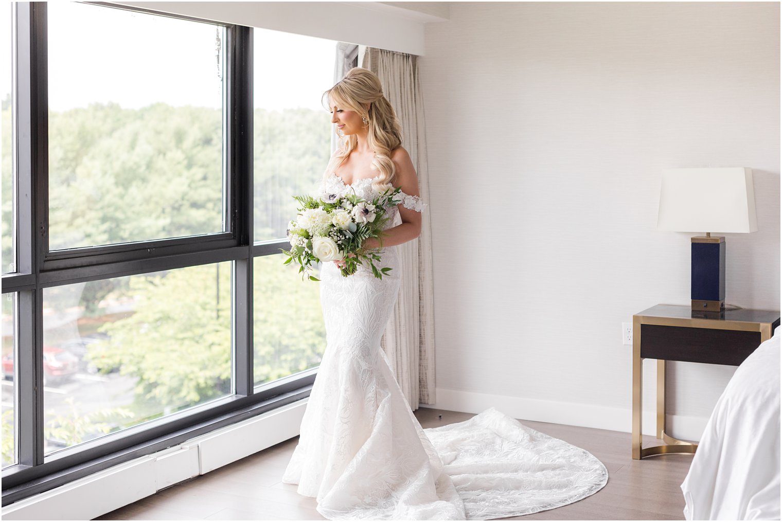 blonde bride looks out window holding ivory bouquet with strapless gown and lace straps 