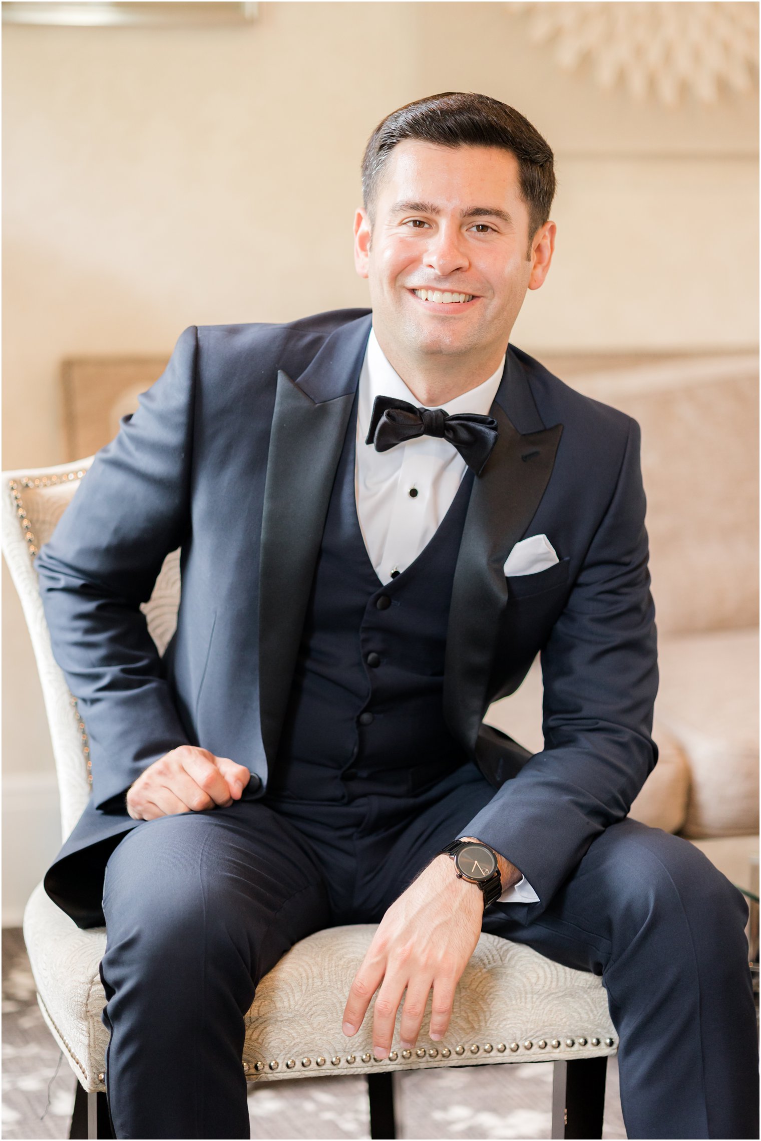 groom in navy suit with black lapels sits on tan chair at The English Manor