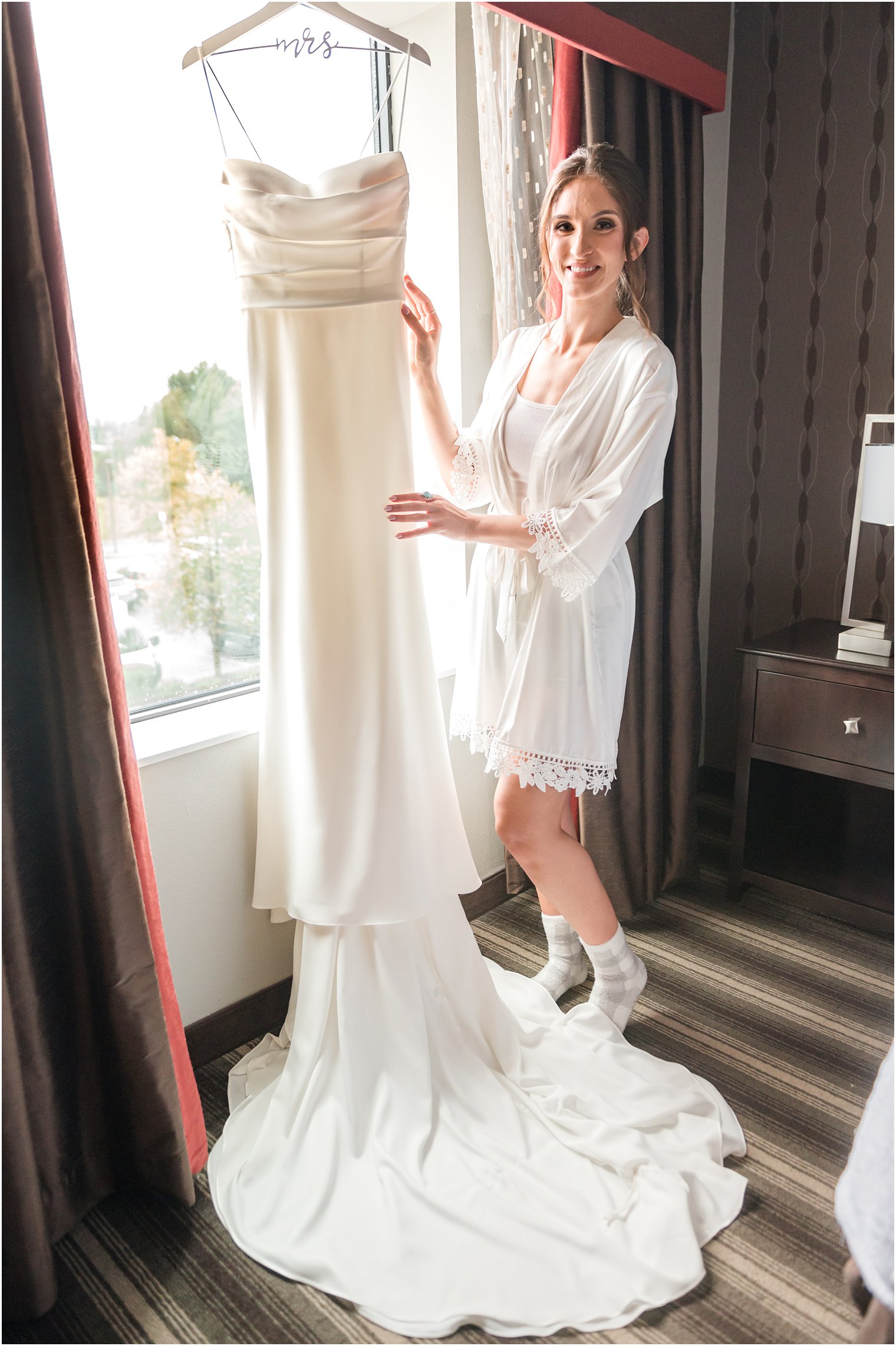 bride in white robe poses by modern wedding gown hanging in window 