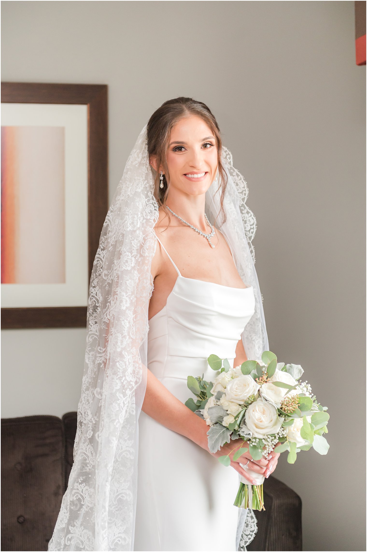 bride stands in sleek wedding gown with thin straps with lace veil around her shoulders 
