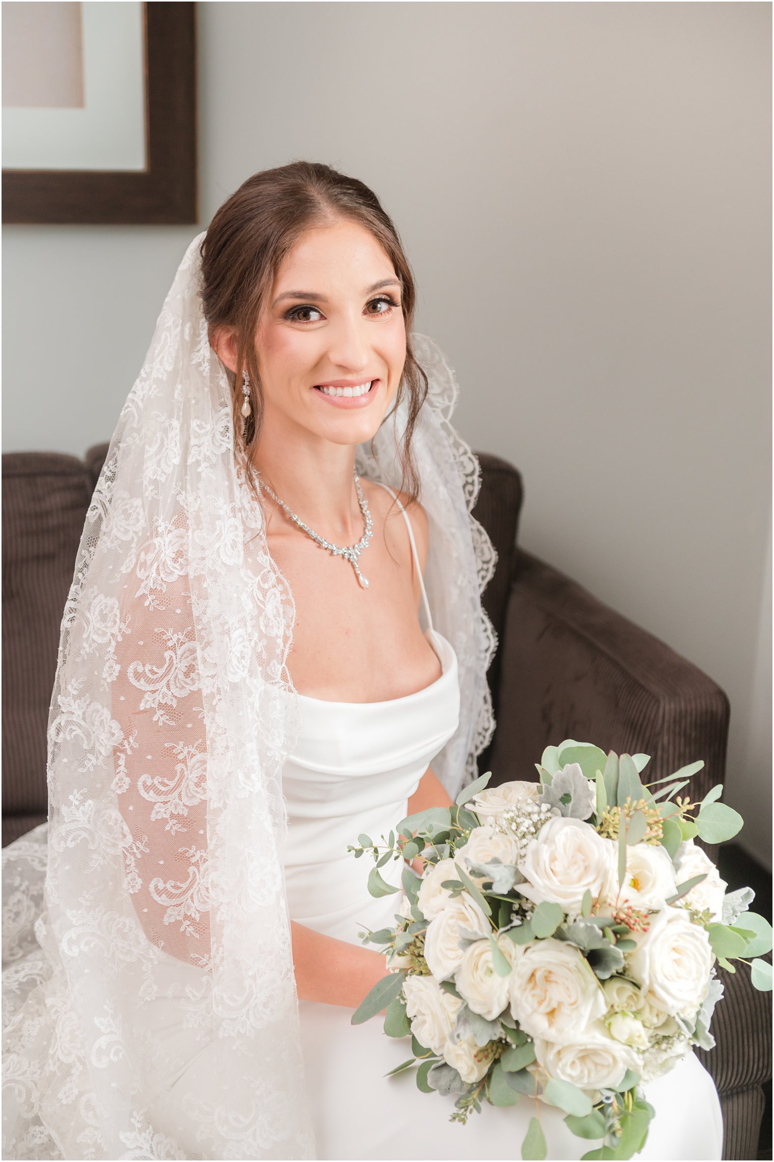 bride in sleek wedding dress with thin straps sits on brown chair with veil around her holding all-white bouquet 