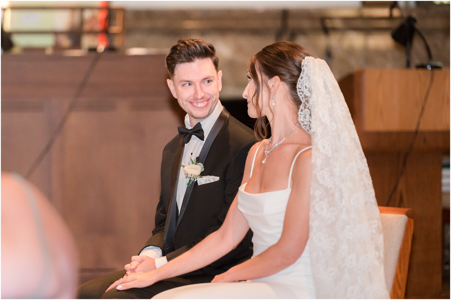 groom smiles at bride sitting together during traditional church ceremony in small New Jersey church