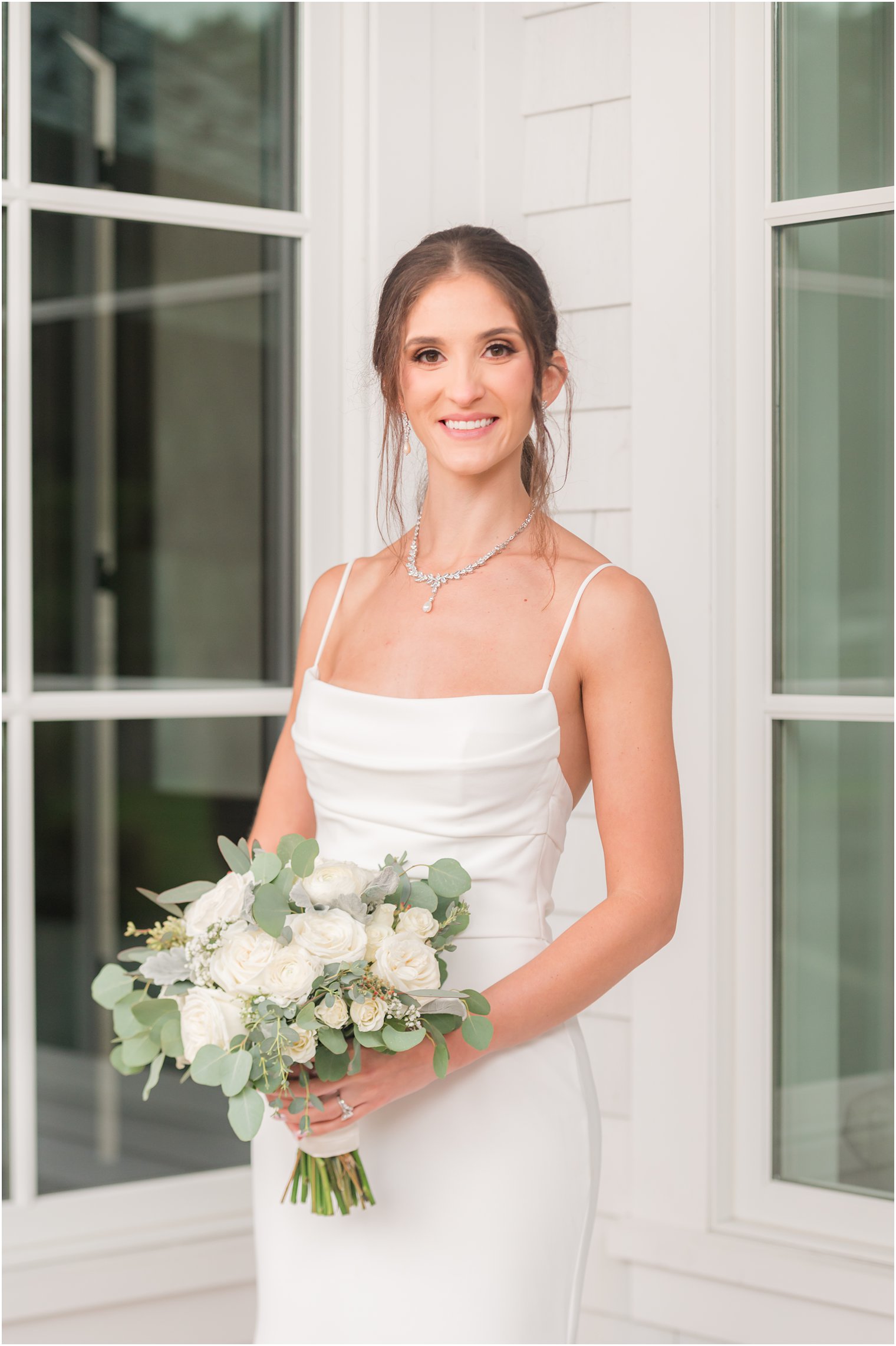 bride in sleek wedding gown holds bouquet of white roses by white wall at The Mill Lakeside Manor