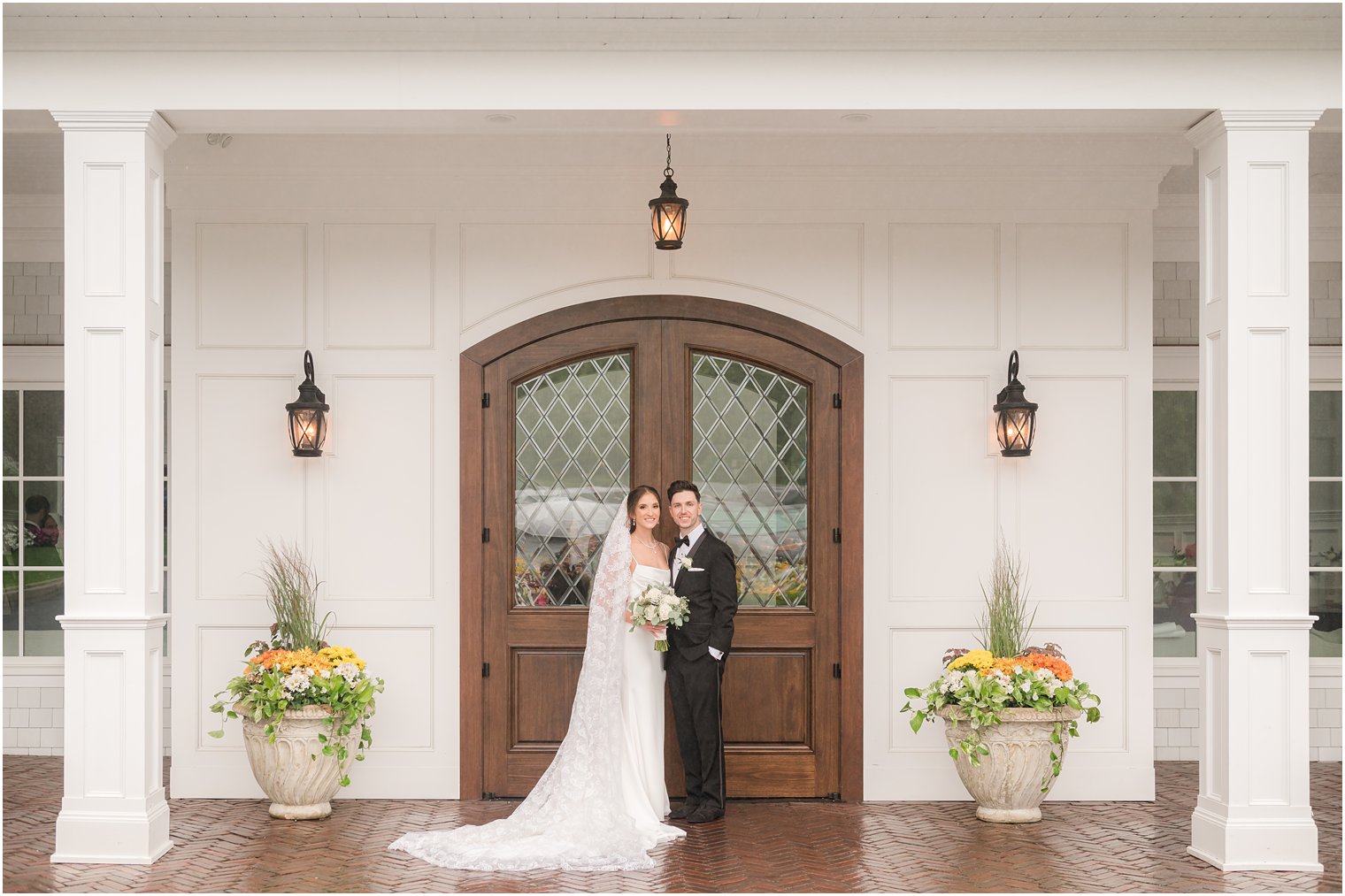 bride and groom stand hugging by wooden door with detailed lace veil down bride's shoulders 