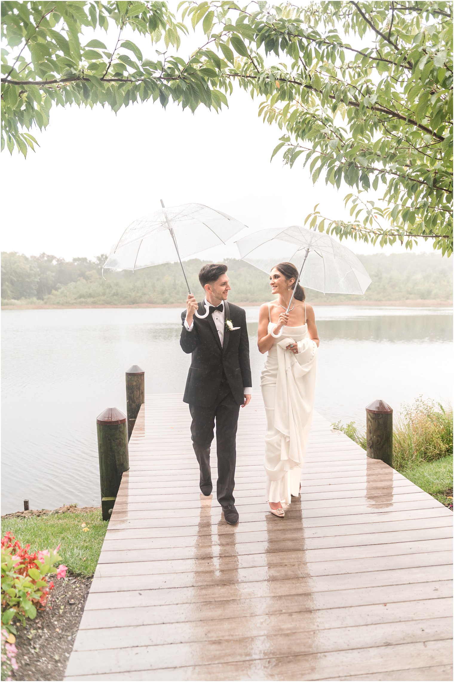 bride and groom walk on wooden dock under clear umbrellas on rainy wedding day at The Mill Lakeside Manor
