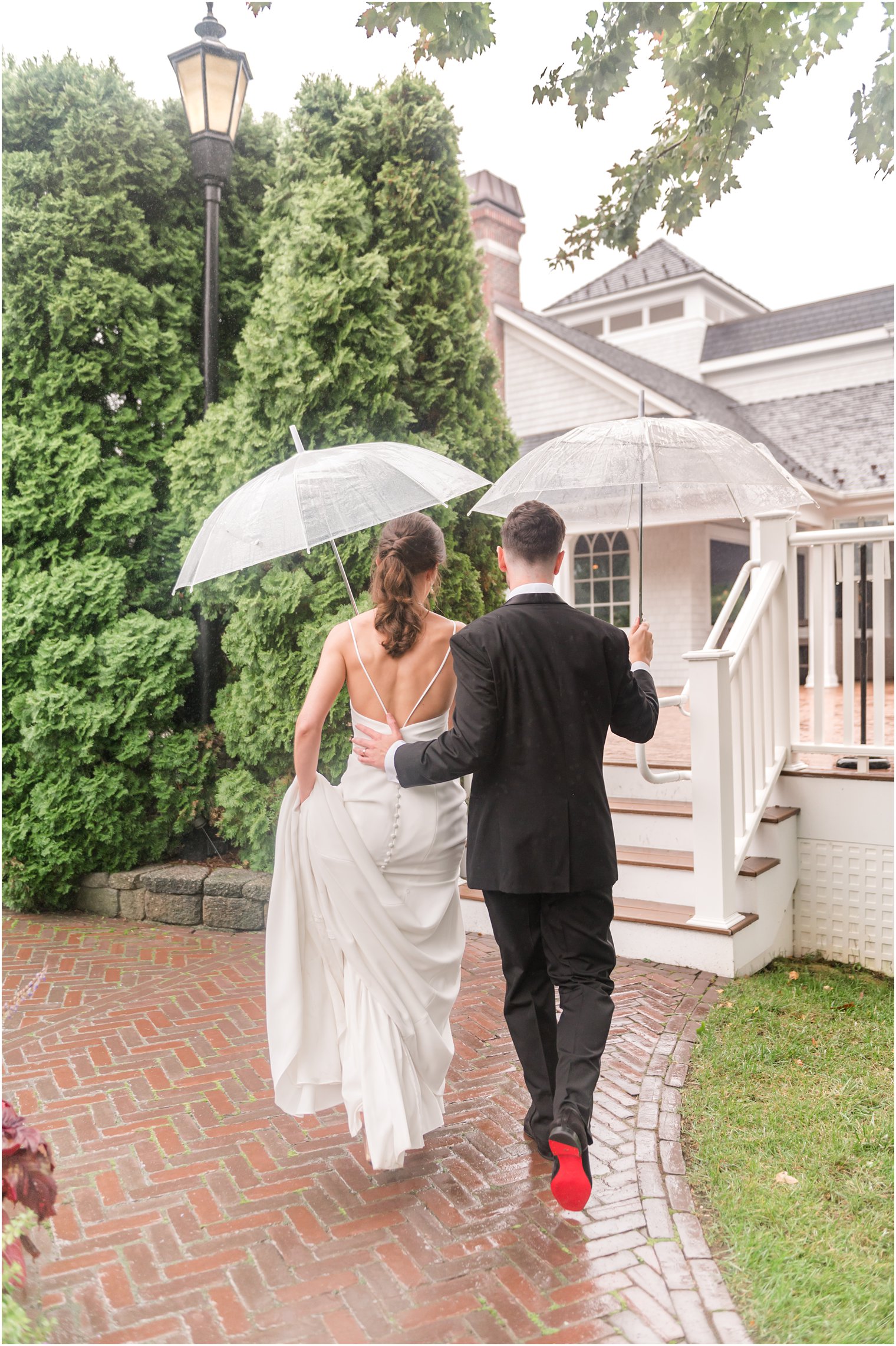 groom walks with bride up to The Mill Lakeside Manor under clear umbrellas on rainy wedding day