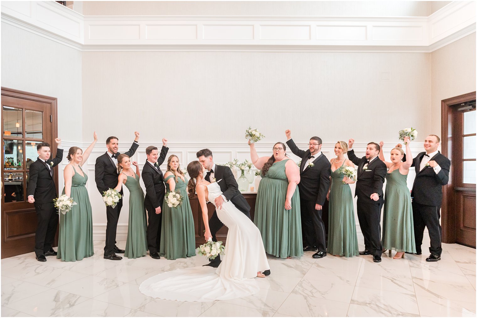 groom dips bride kissing her while wedding party in black and sage green cheer