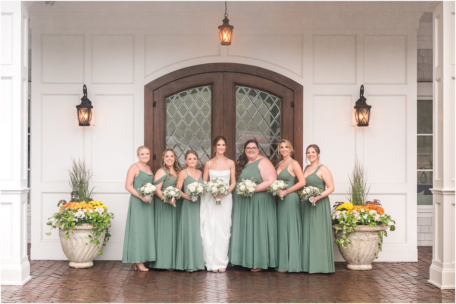 bride poses with bridesmaids in sage green gowns outside wooden door at The Mill Lakeside Manor