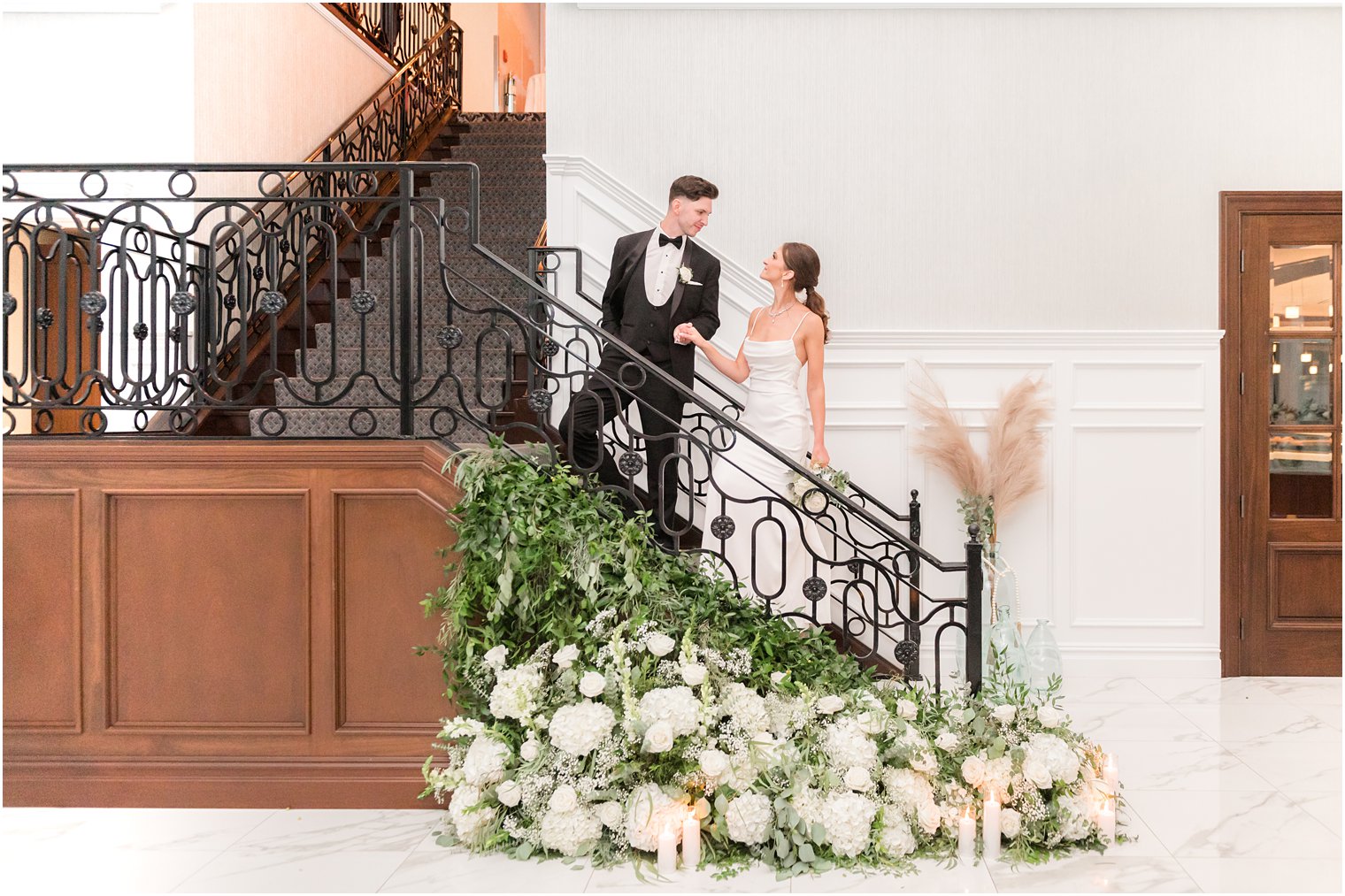 groom leads bride up stairs with white and green floral display 