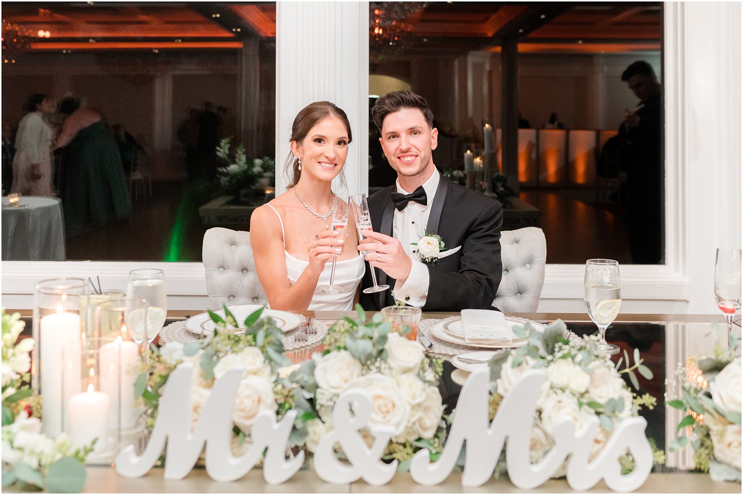 newlyweds sit together at sweetheart table with white Mr and Mrs sign and white roses 