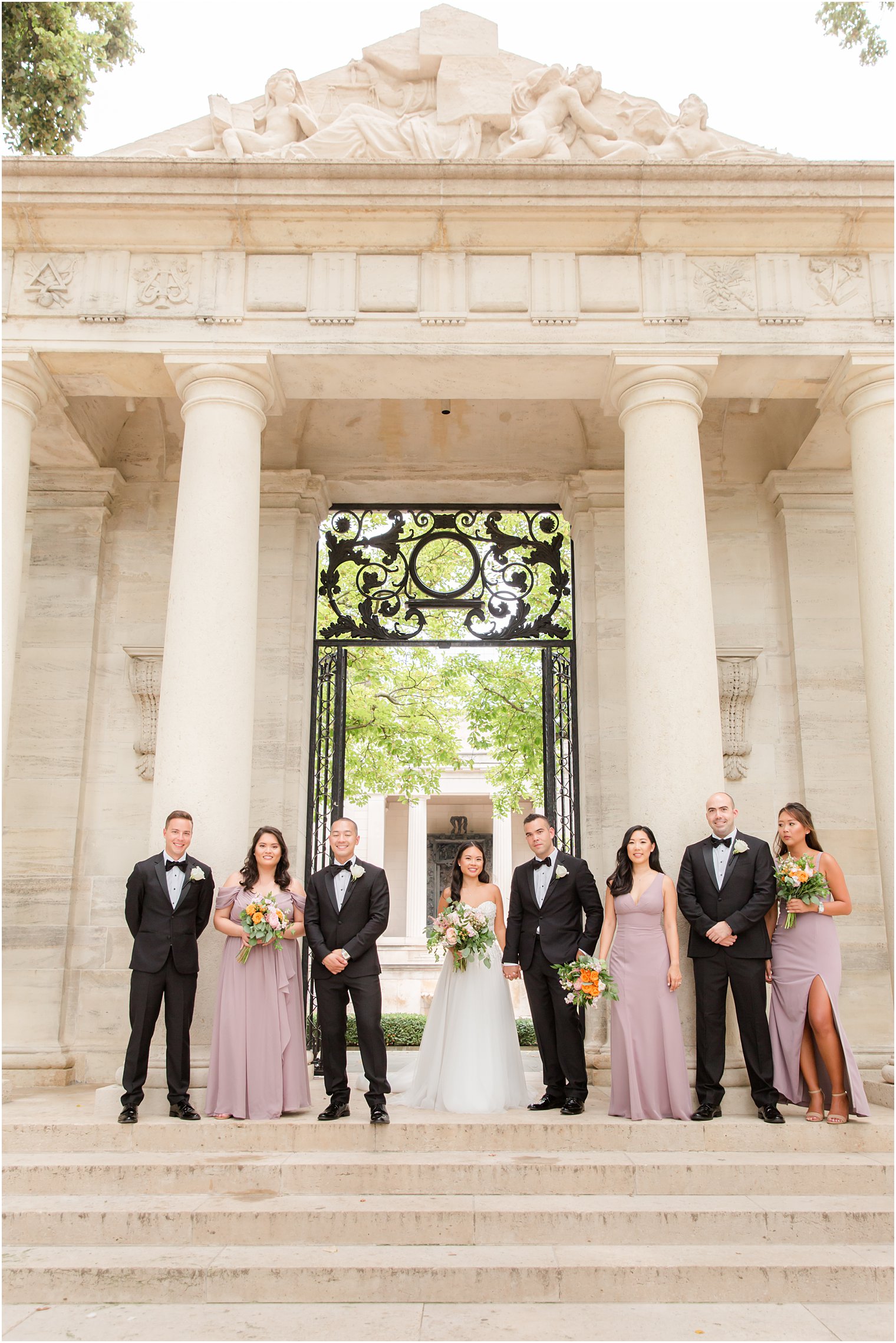 newlyweds stand with wedding party in black tuxes and mauve gowns on steps of the Rodin Museum in front of columns 