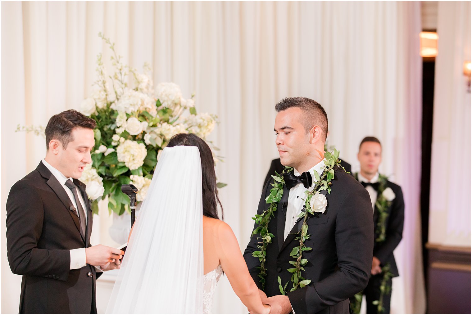 groom with greenery around neck smiles at bride during ceremony at the Ballroom at the Ben