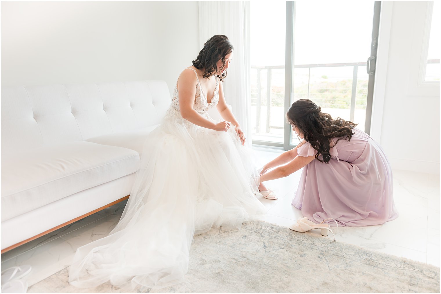 bridesmaid in pastel purple gown helps bride with shoes 
