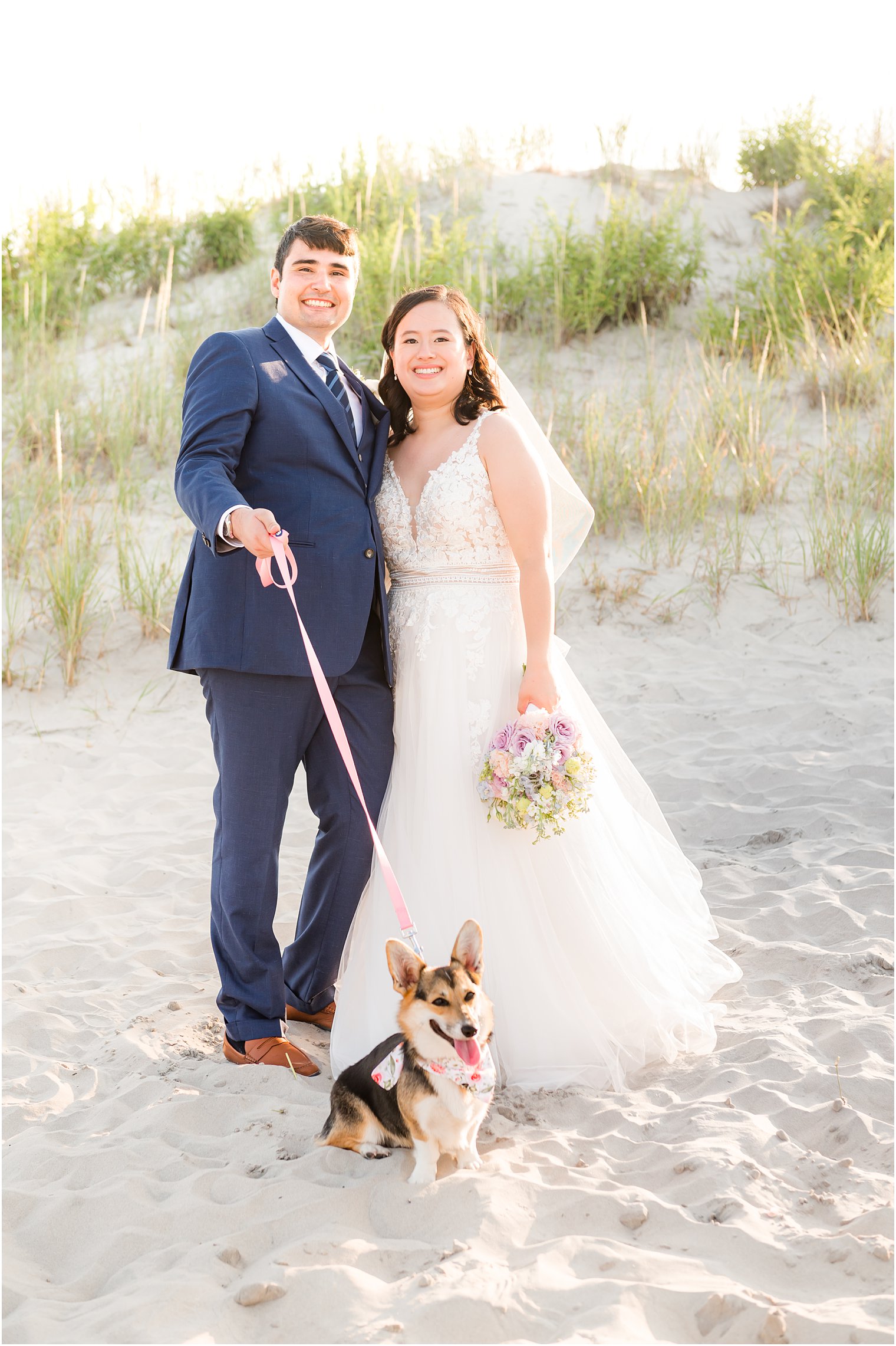 bride and groom stand on beach with dog on leash in front of them
