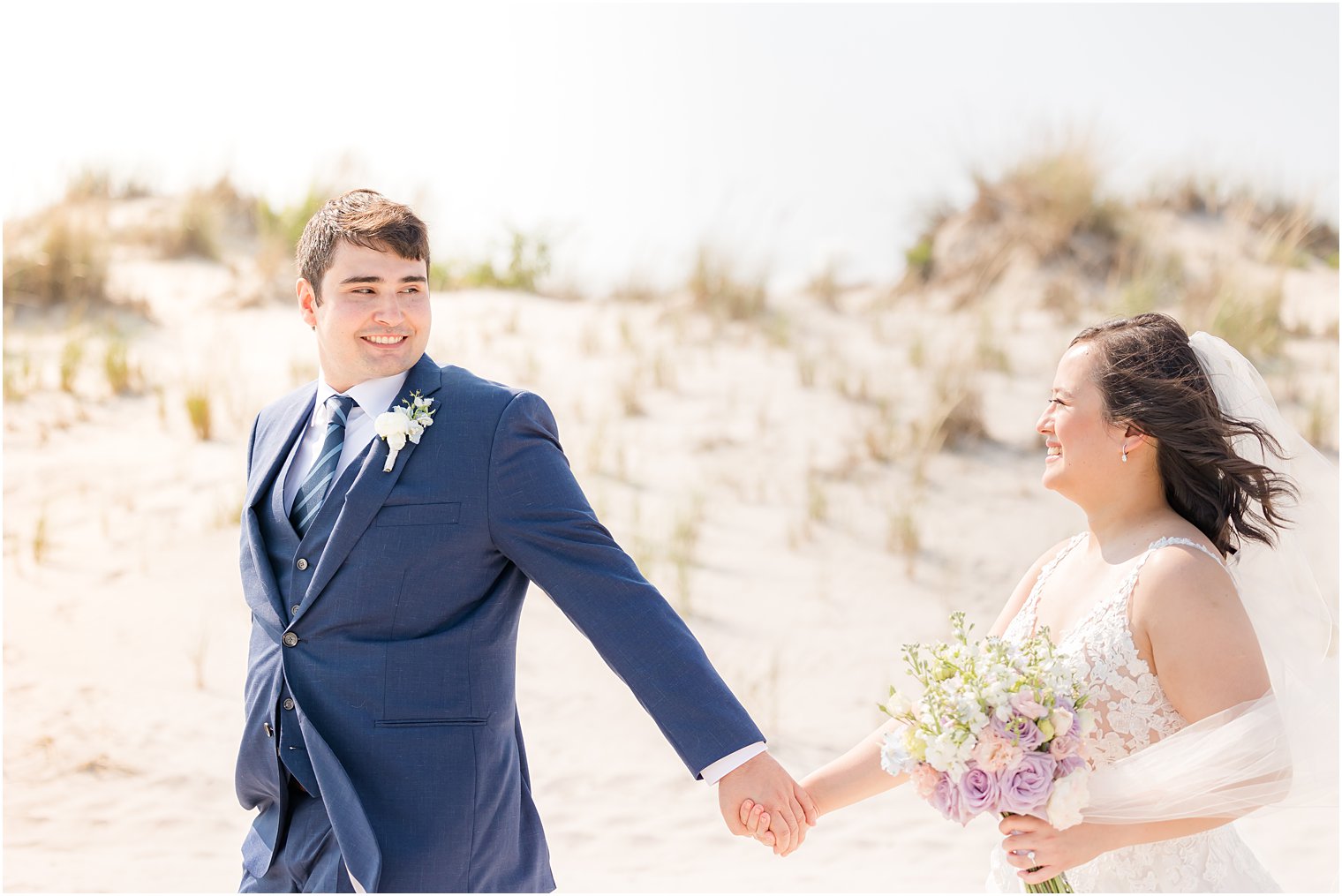 groom in navy suit leads bride across beach in Avalon NJ