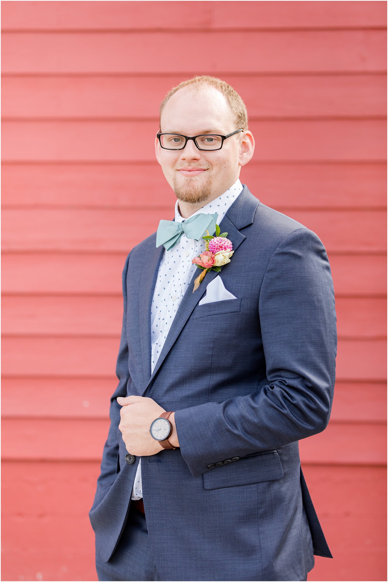 groom adjusts navy blue suit jacket in front of red barn wall 