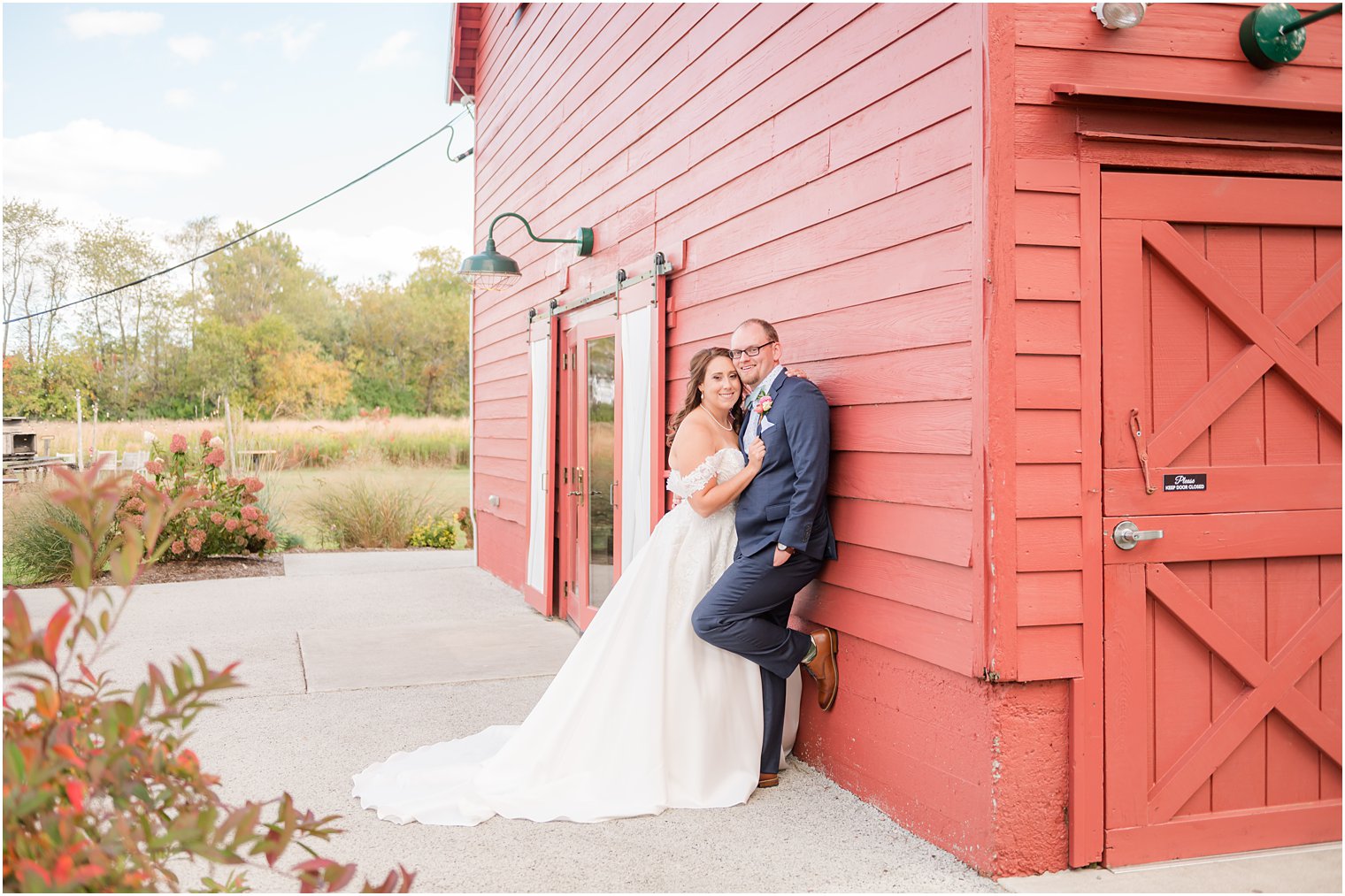 groom leans against wall of red barn at Bishop Hall Farmstead