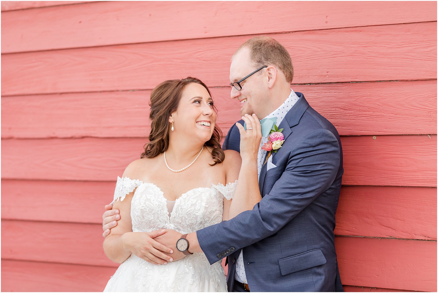 bride reaches up to groom's chin leaning against wall of red barn at Bishop Hall Farmstead