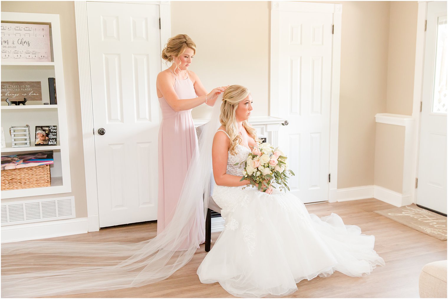 bridesmaid in pink gown helps bride place veil at home 