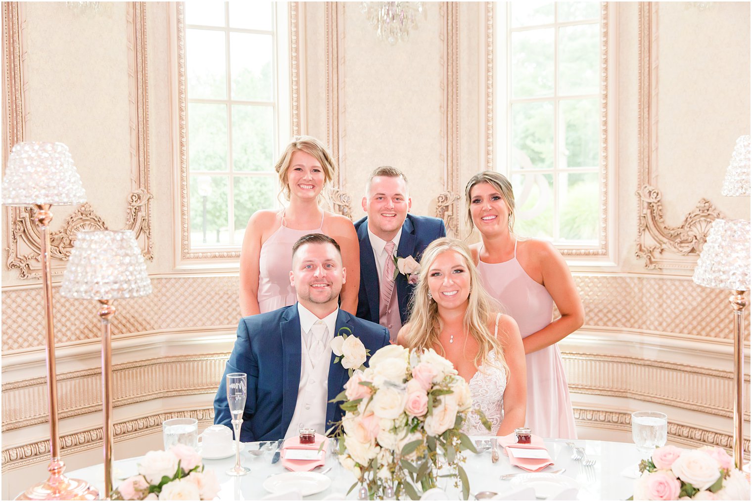 bride and groom sit at table with maids of honor and best man behind them 