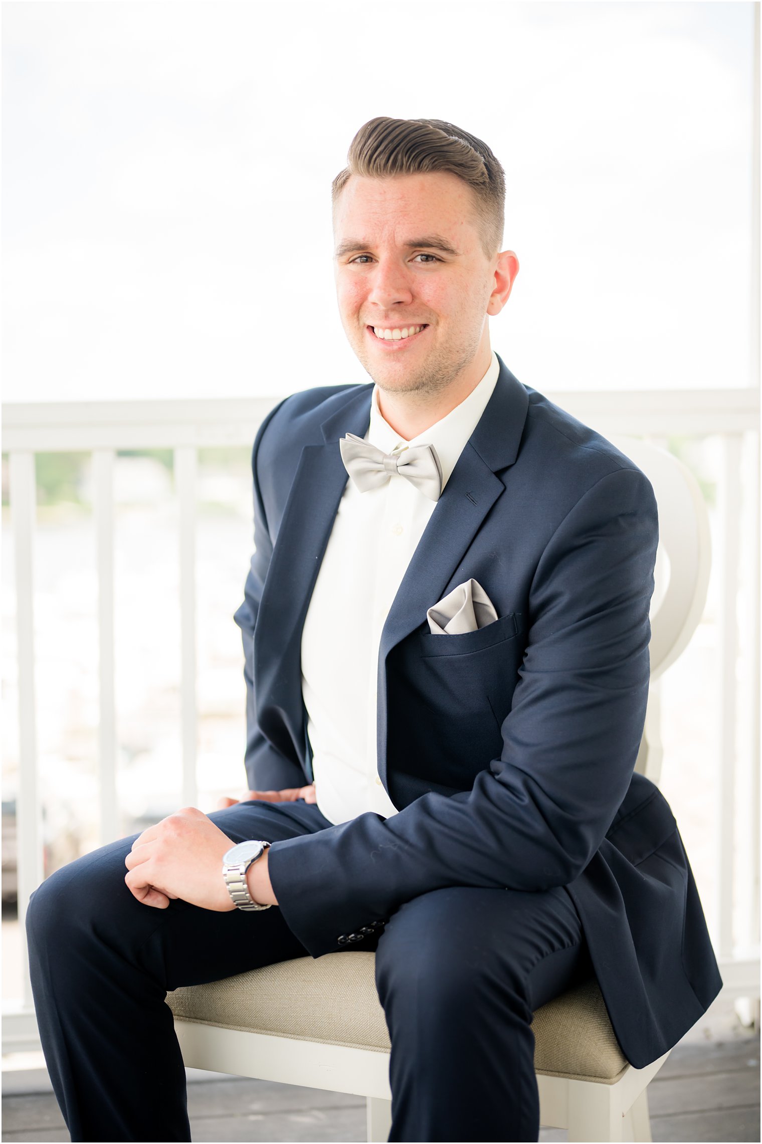 groom sits in navy suit with bowtie on balcony at Crystal Point Yacht Club