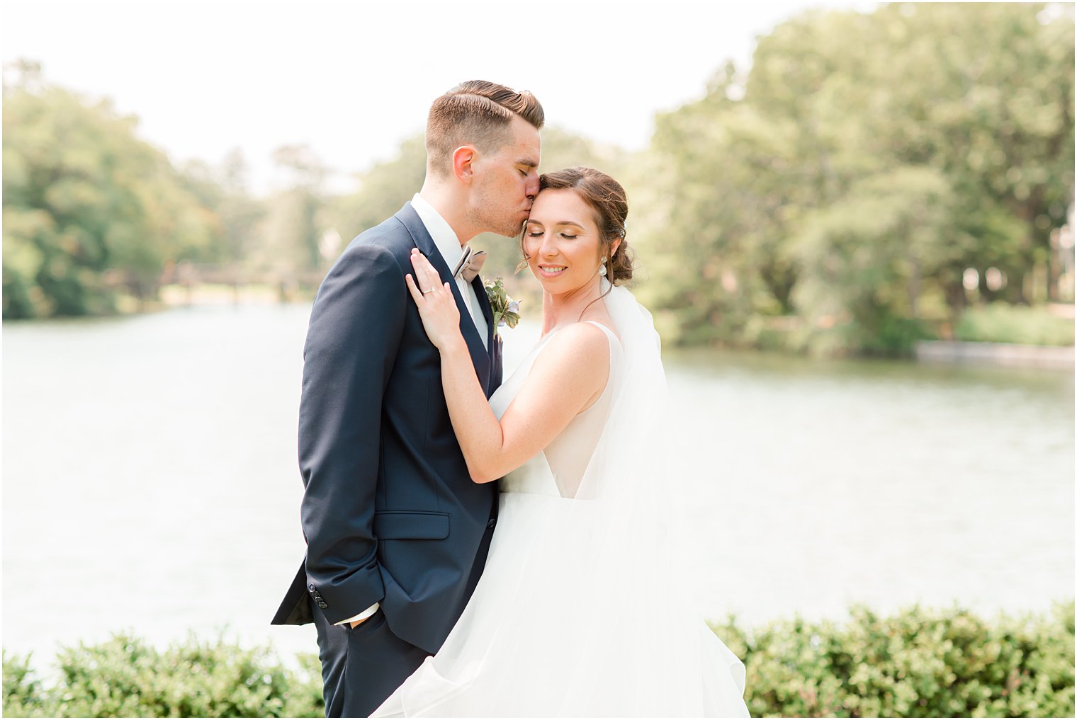 groom kisses bride's head by lake in Spring Lake Park