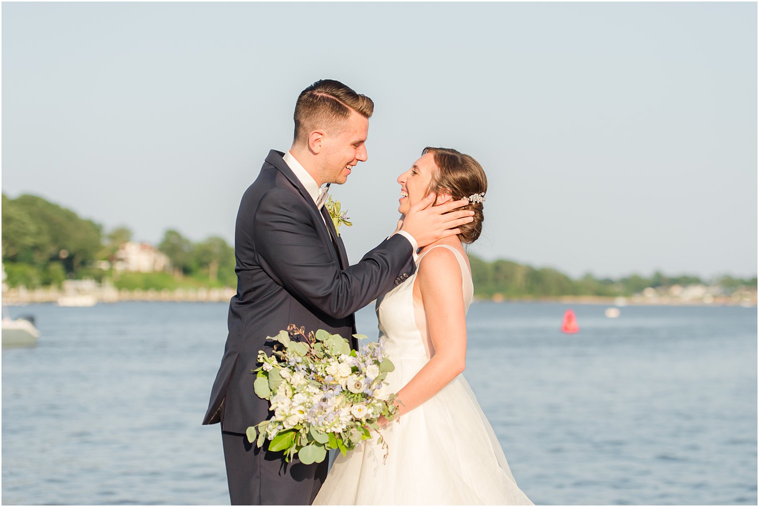 groom holds bride's head leaning for a kiss in front of water