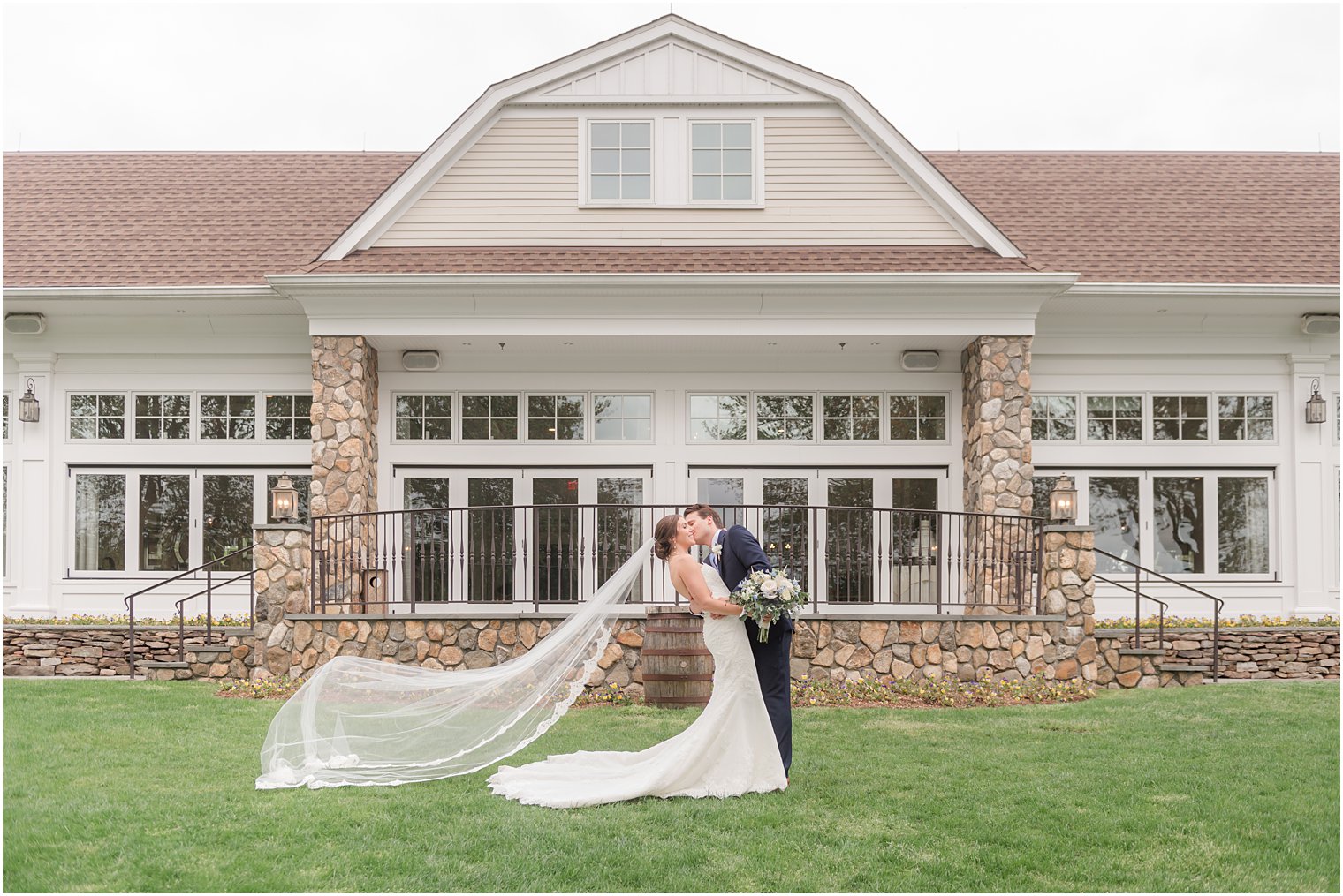 newlyweds kiss with bride's veil floating behind them in front of brick patio at Indian Trail Club