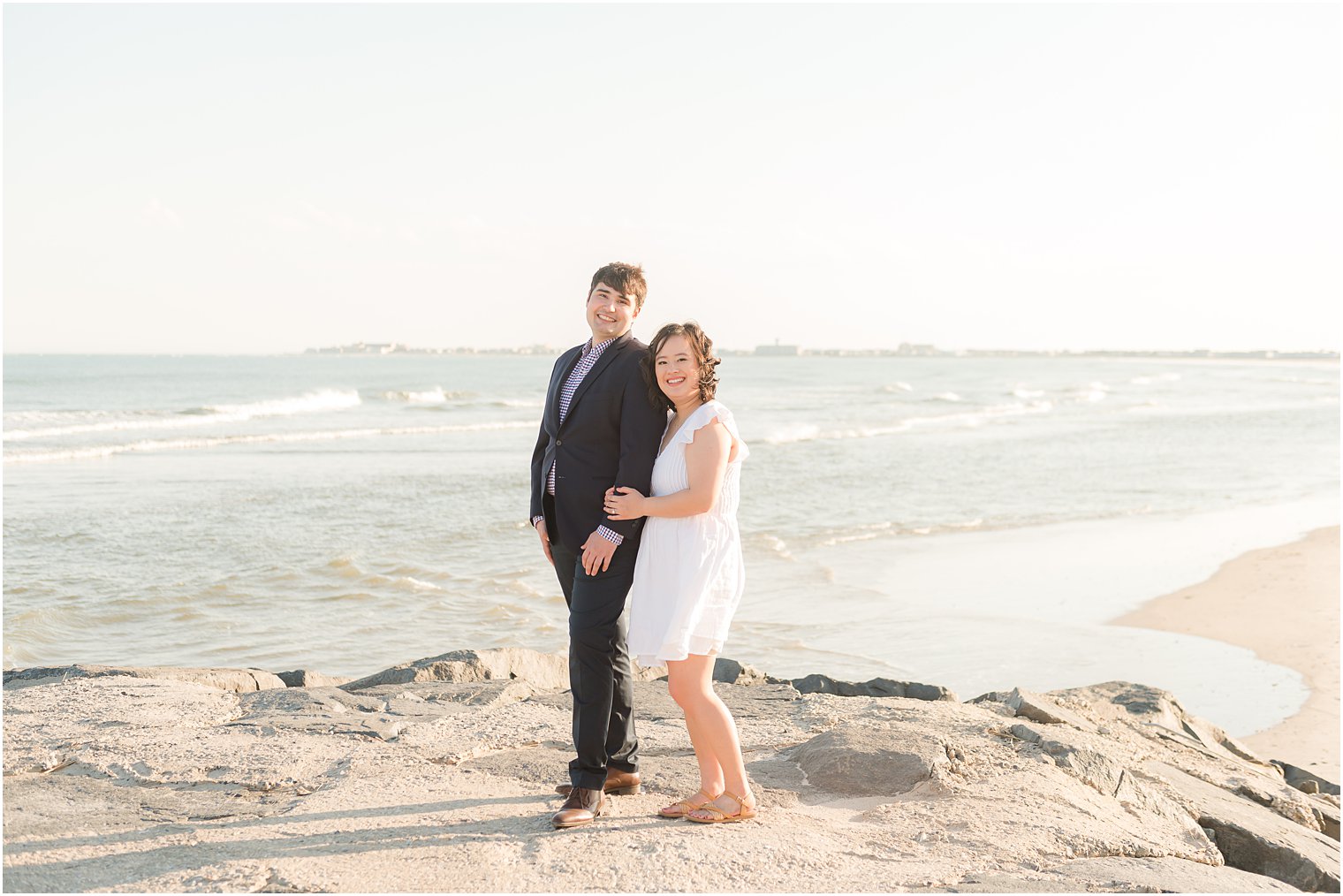 bride hugs groom from behind on rocks during beach engagement session in Avalon NJ