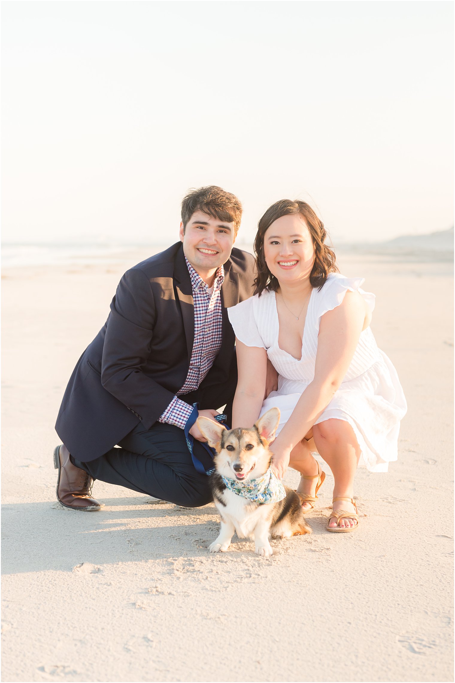 couple kneels behind small dog on beach during engagement photos 