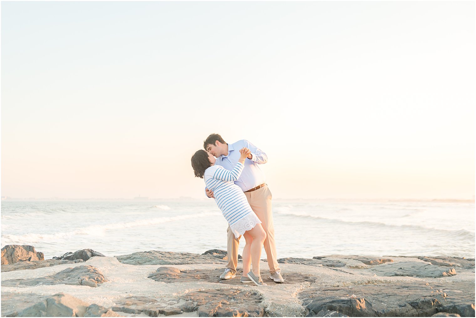 groom kisses bride dipping her while standing on rocks during beach engagement session in Avalon NJ