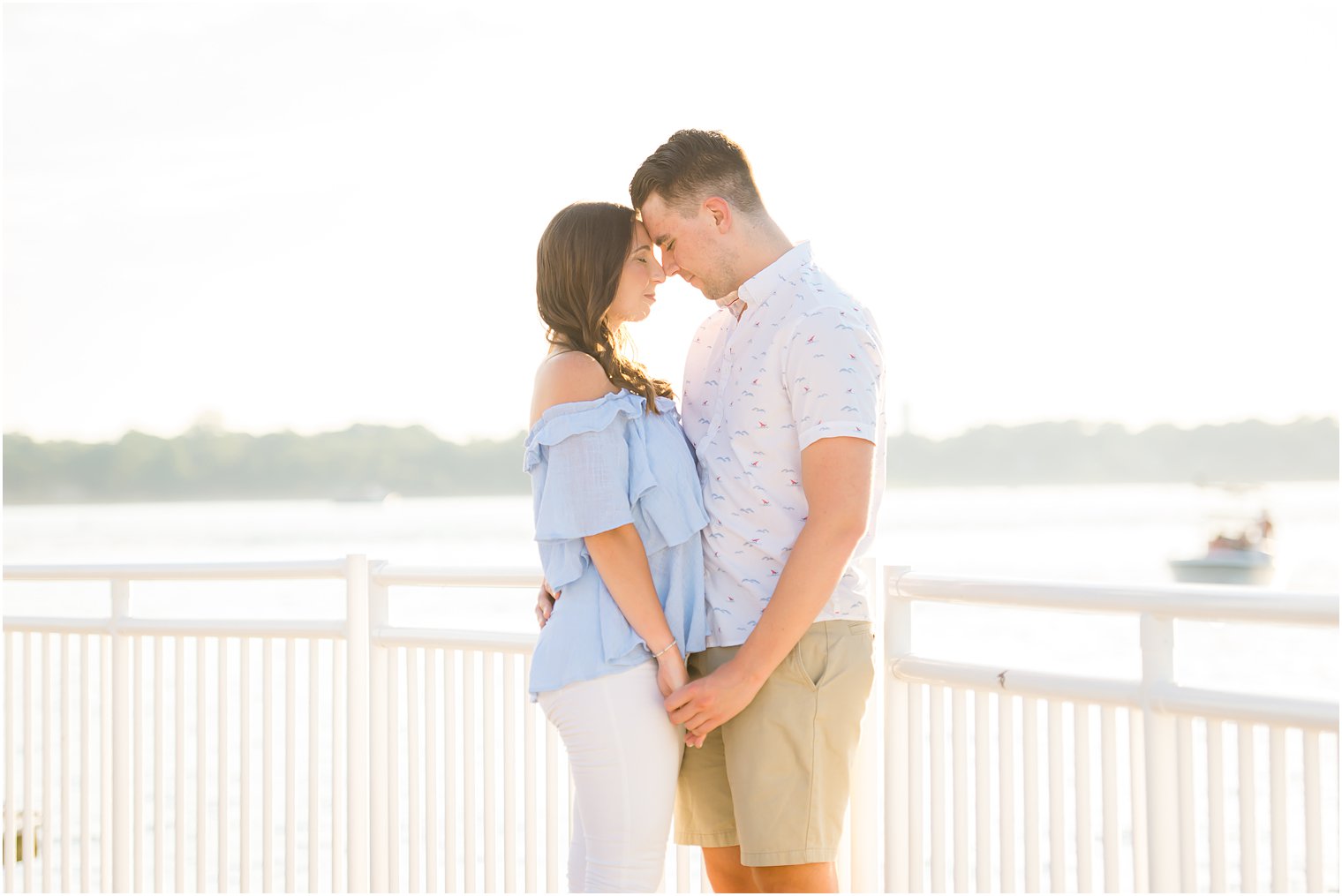 engaged couple stands together against white picket fence with heads touching 