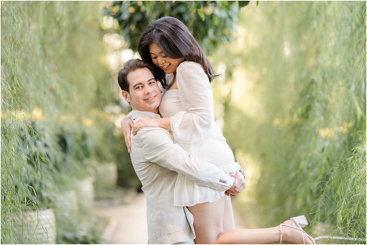 man lifts up fiancee among weeping willows at Longwood Gardens