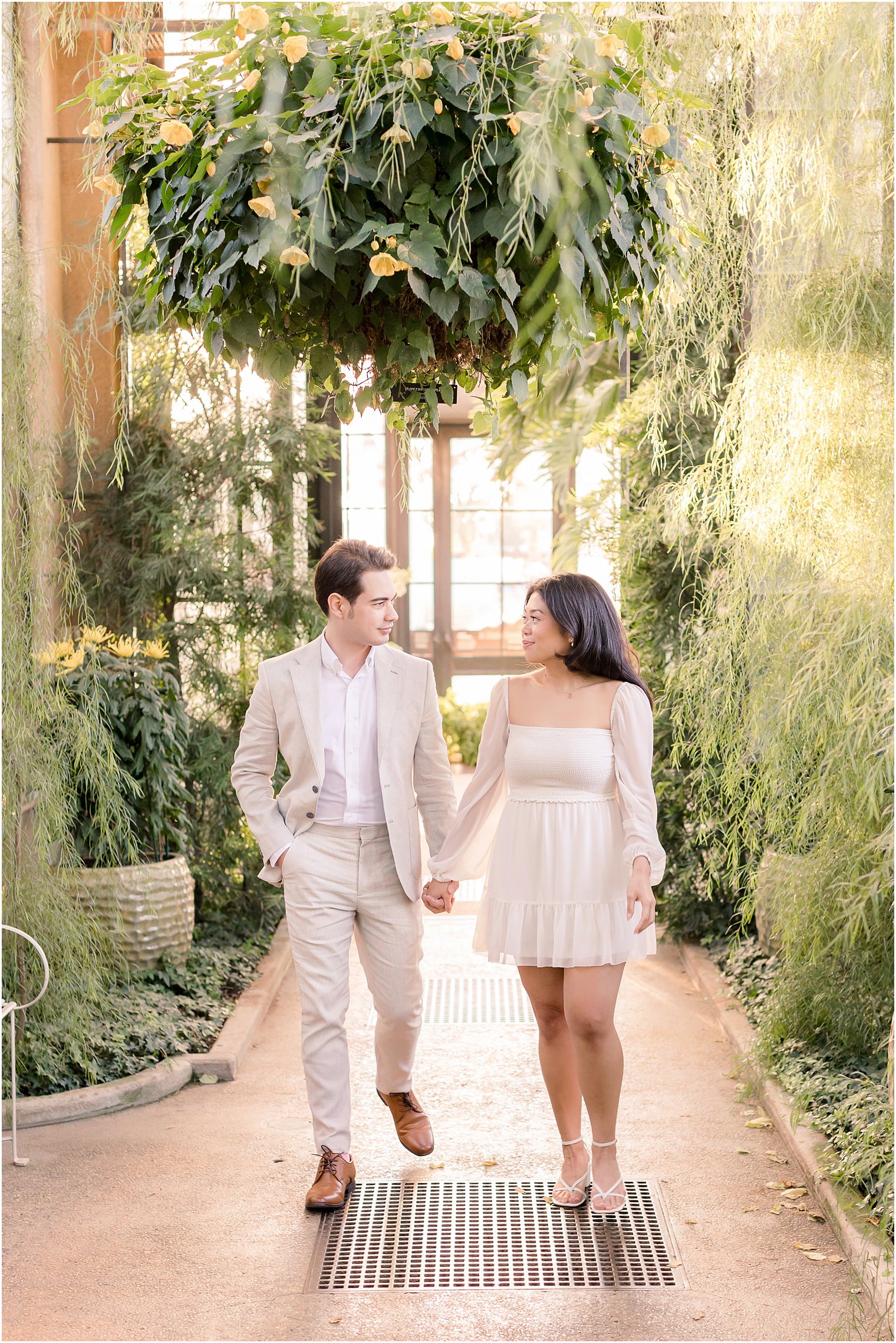 engaged couple holds hands walking under hanging plants 
