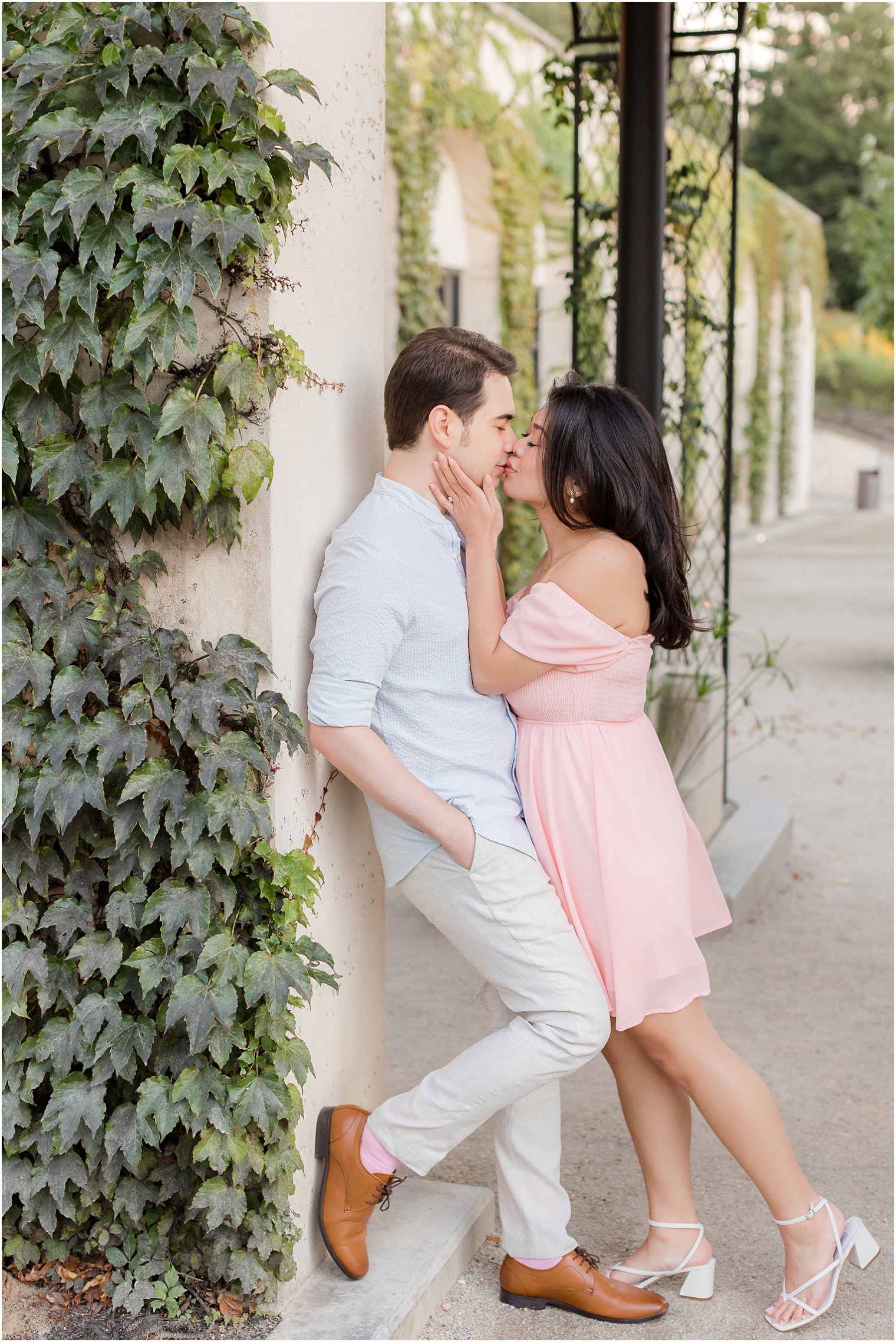 man in blue shirt leans against wall while woman in pink dress leans to kiss him