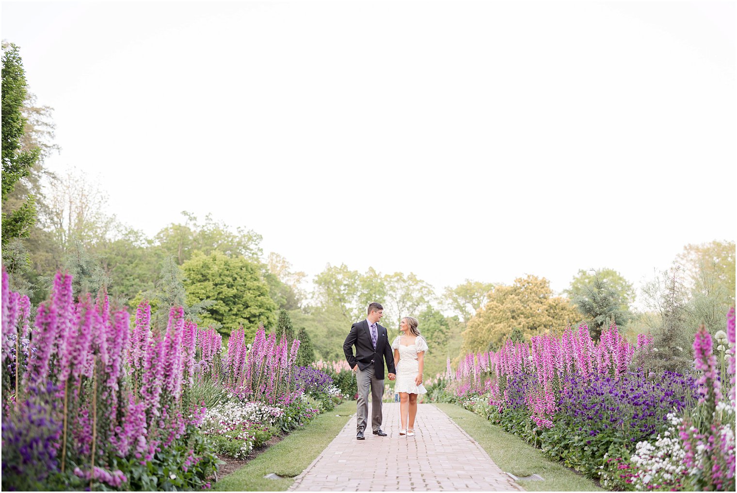 engaged couple holds hands waking through pink and purple flowers during Longwood Gardens engagement session