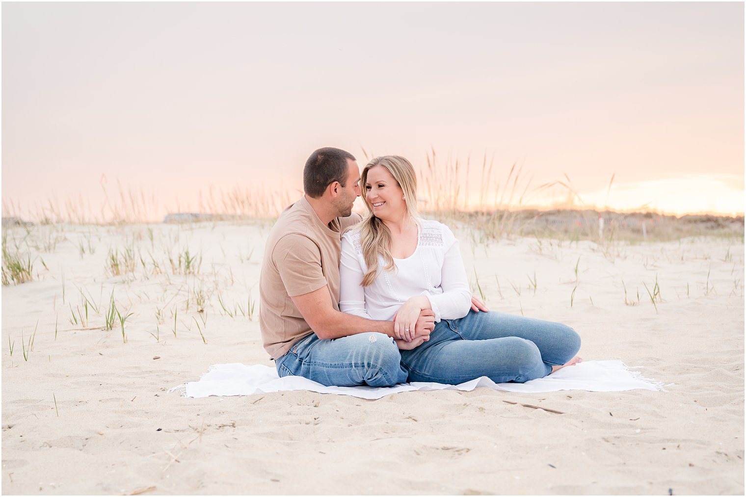 engaged couple sits on white blanket at sunset at Spring Lake Beach