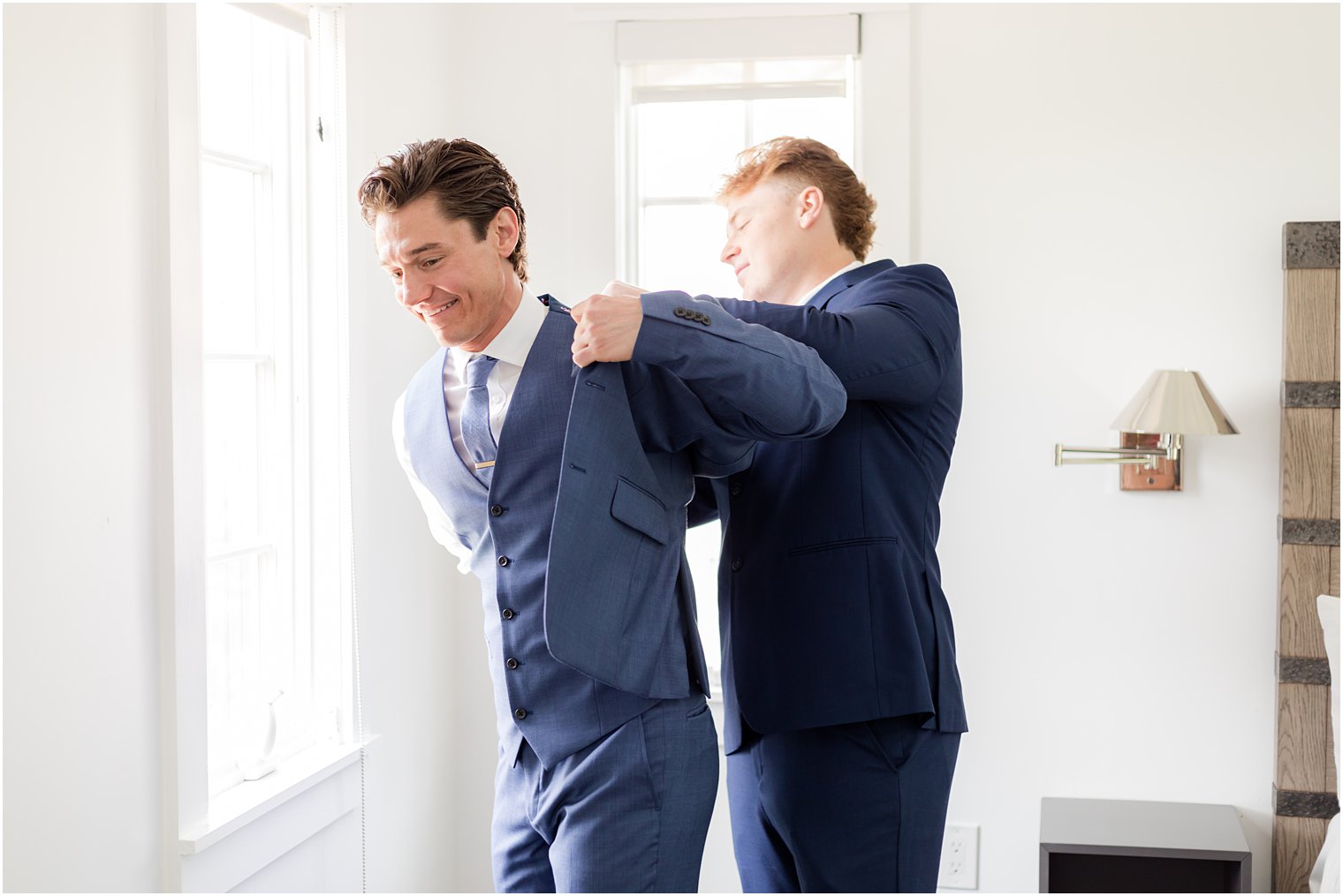 groomsman helps groom into navy suit jacket in suite at Ryland Inn