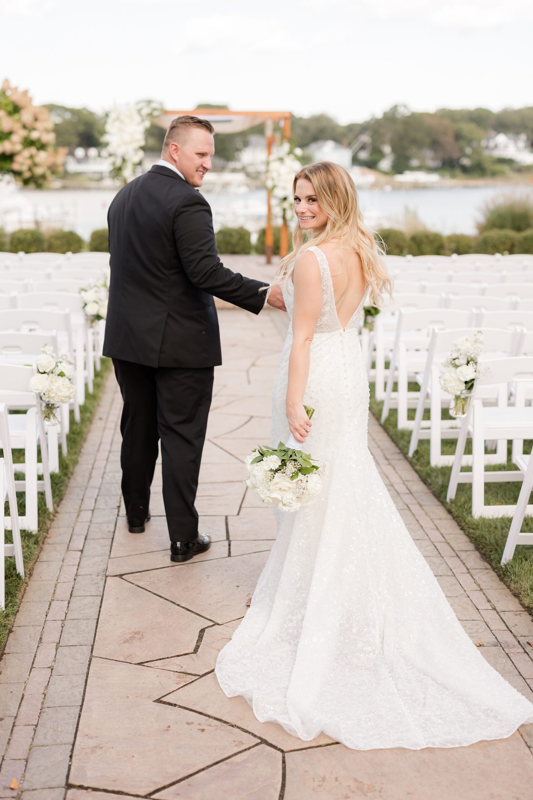 Bride and groom walk down an outdoor aisle, glancing back and smiling, with waterfront and greenery in the background