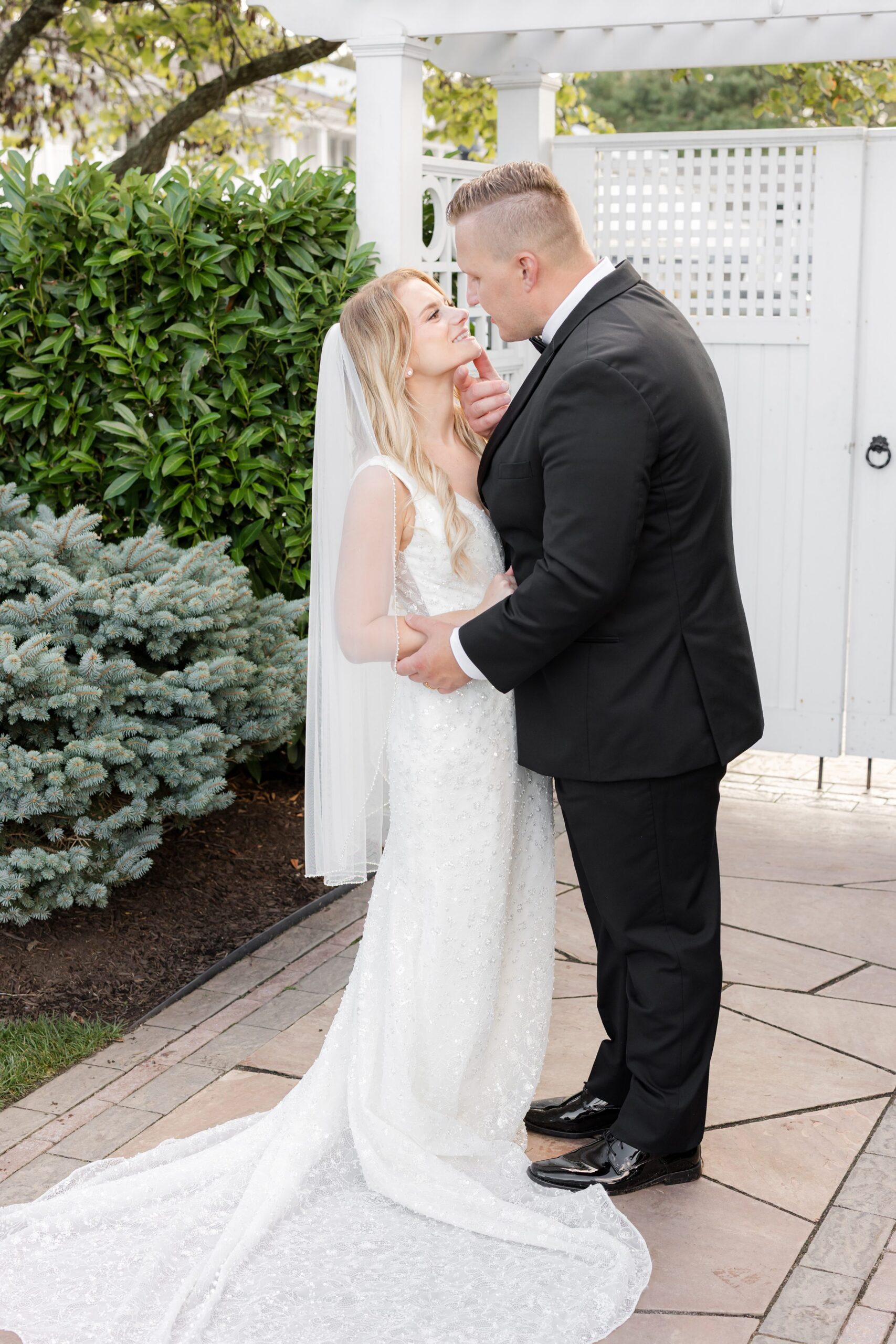 Bride and groom stand closely together outdoors, smiling at each other, framed by greenery and a white garden arch in a romantic, intimate moment